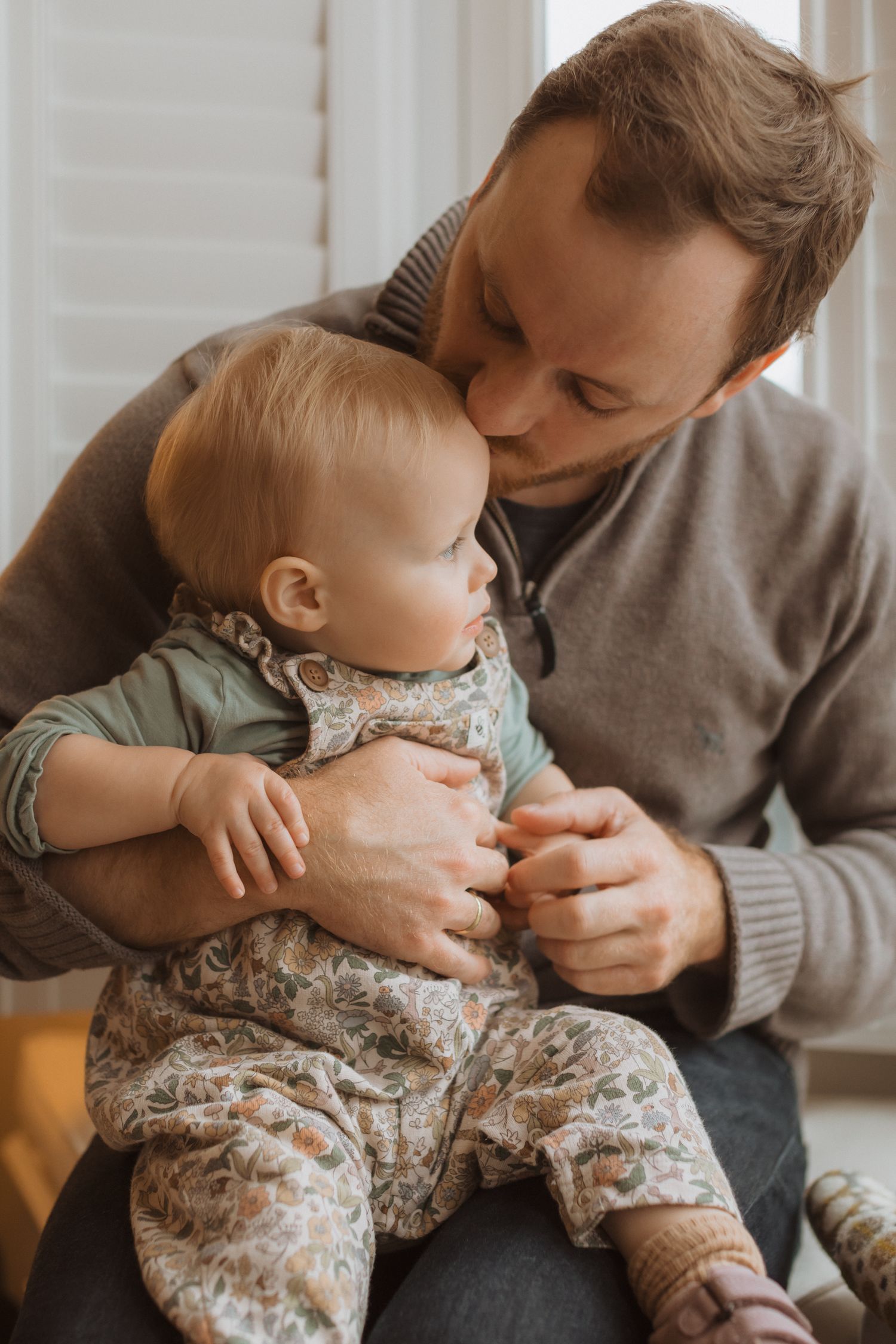 Tender parenting moment showing gentle interaction between adult and baby in floral outfit near window.