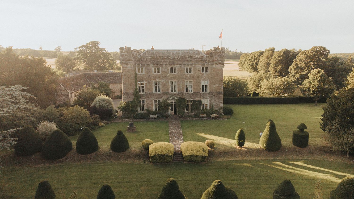 A scenic panorama of a historic manor house with manicured lawns and gardens captured in warm evening light.