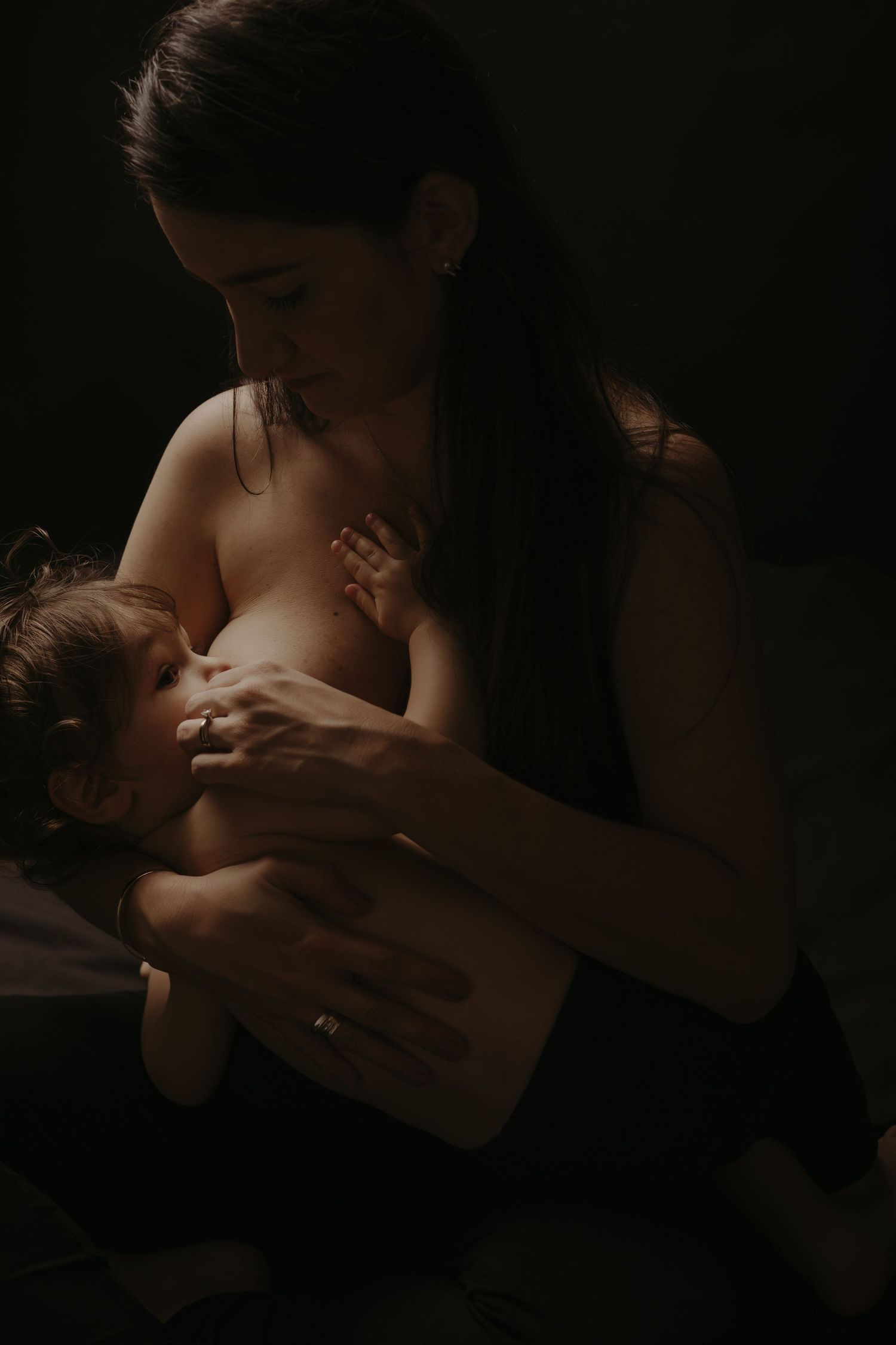 A tender moment of breastfeeding captured in dramatic low-key lighting against a dark background.