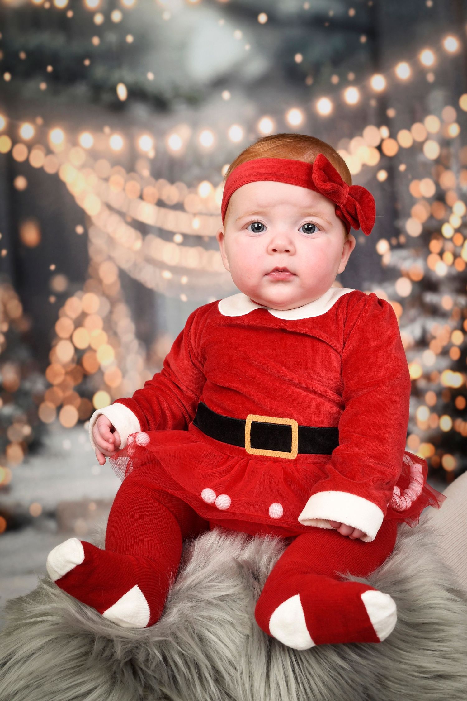 A baby wearing a Santa outfit is seated on a cozy furry blanket, featured in a Christmas photography session at Abigails studio.