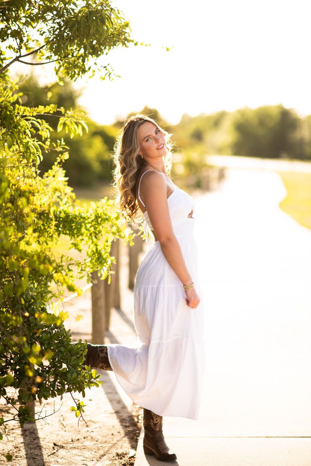 A person in a flowing white dress poses gracefully by a lake at golden hour with sunlit trees in the background.