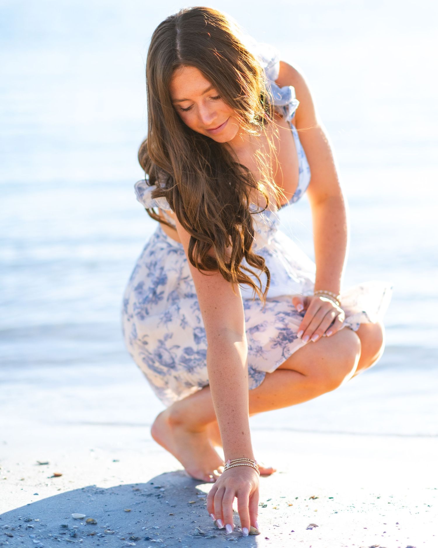 A person in a floral summer dress crouches down on a sunny beach, touching the sand with one hand.