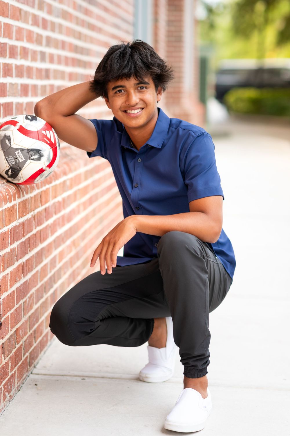 Soccer athlete in navy blue polo and black joggers crouches against brick wall holding red and white ball.