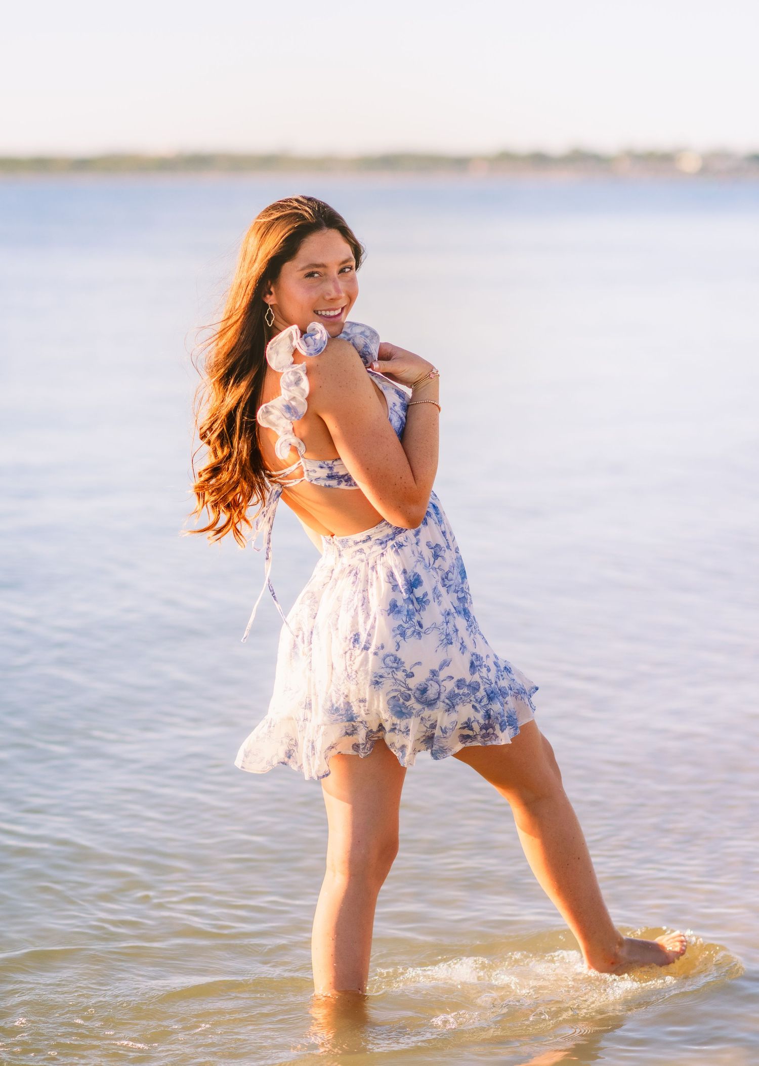 Woman in white and blue floral dress walks barefoot in shallow water at sunset.