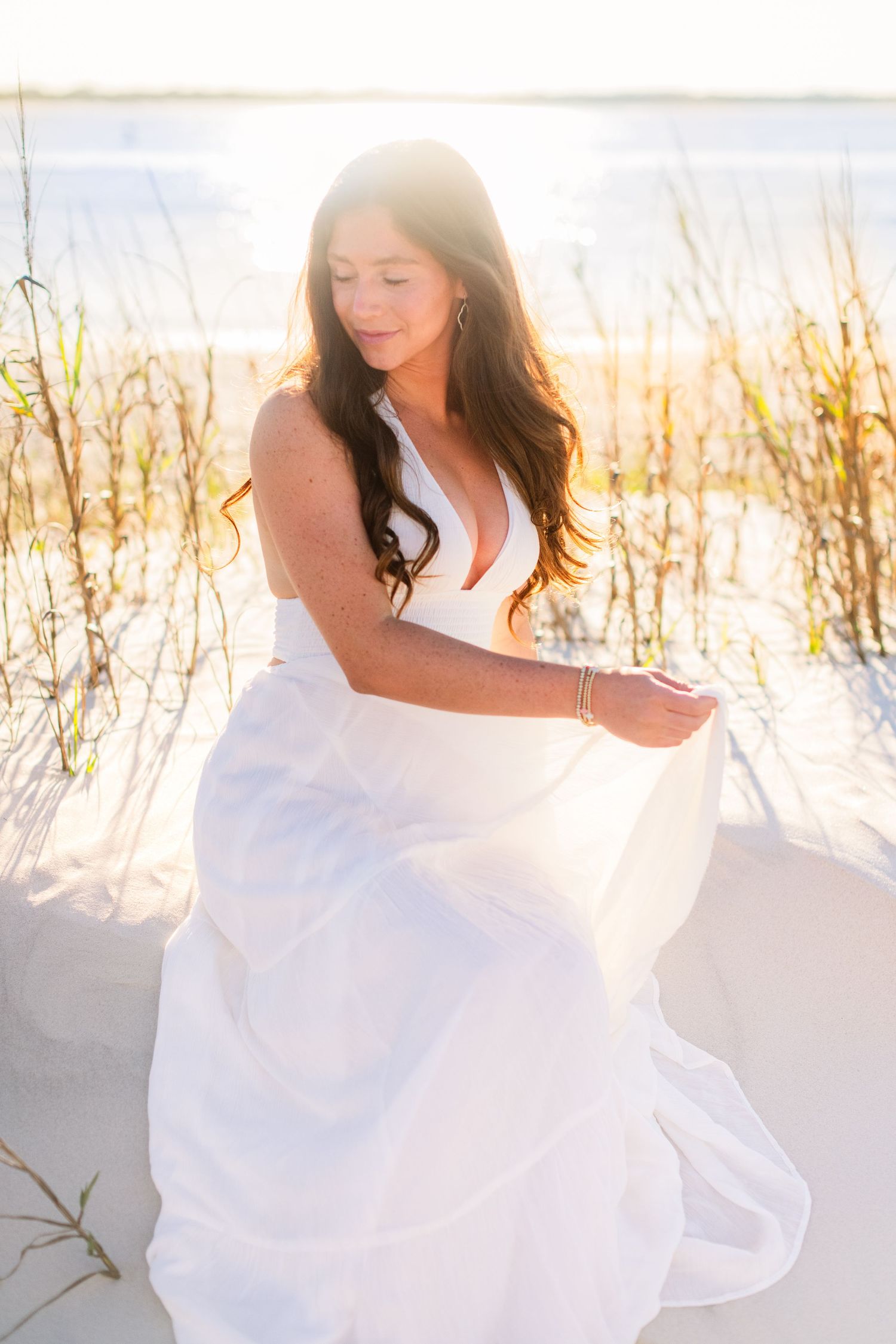 Dreamy beach photoshoot with flowing white dress in coastal grass during golden hour sunset.