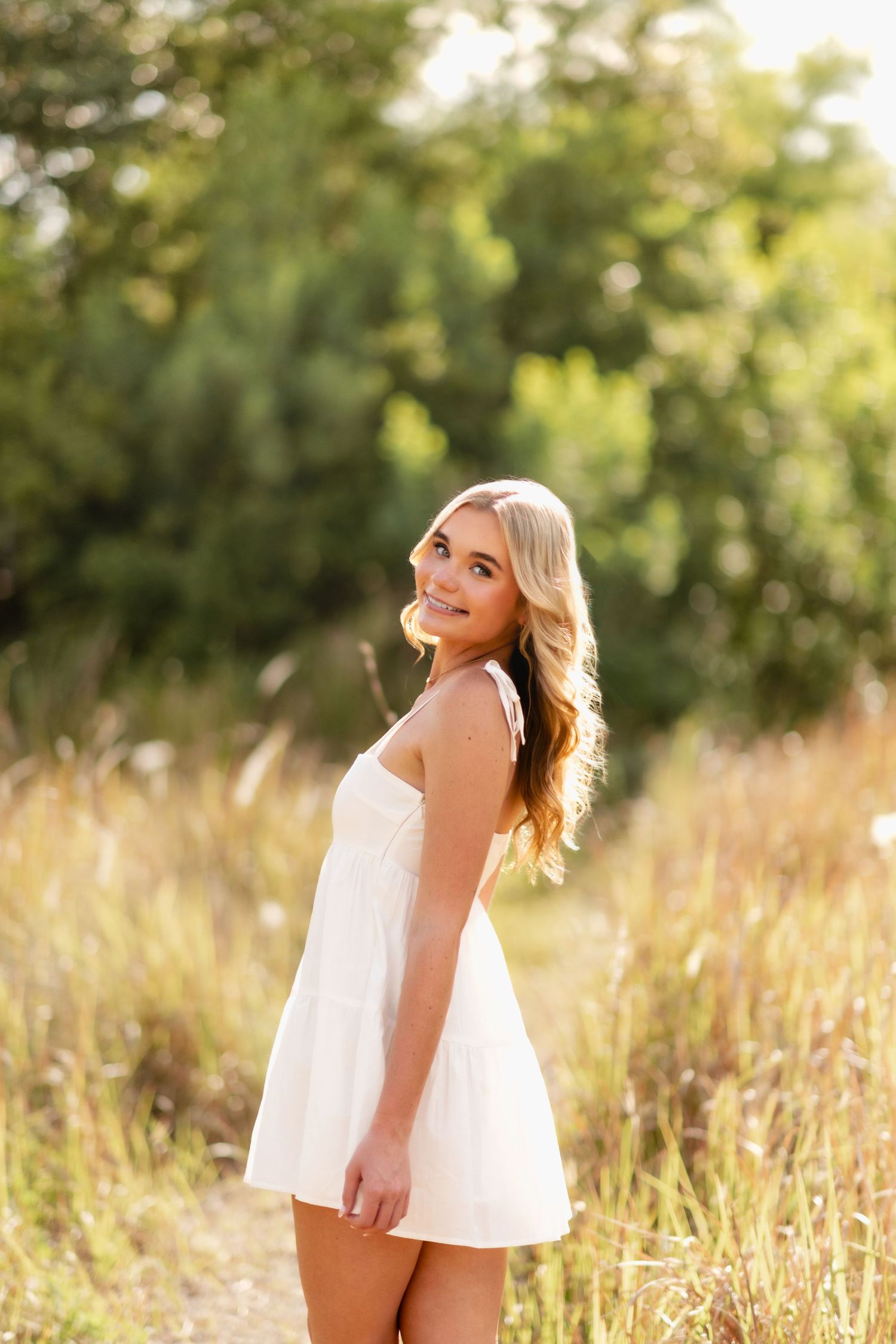 Young blonde woman in white sundress smiling outdoors in golden field with green trees in background.