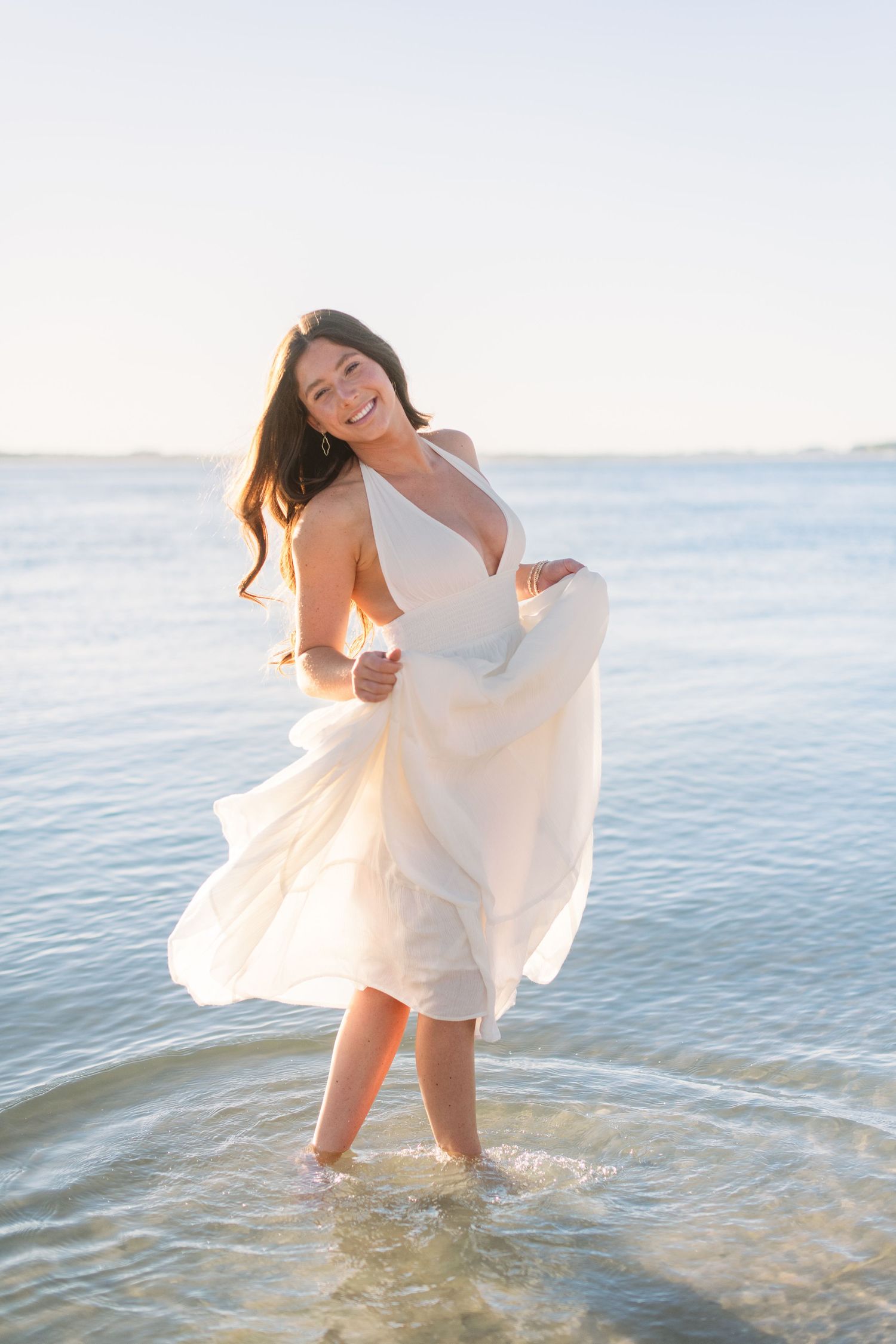 A person in a flowing white dress dances joyfully in shallow ocean waters at sunset, creating a dreamy seaside scene.