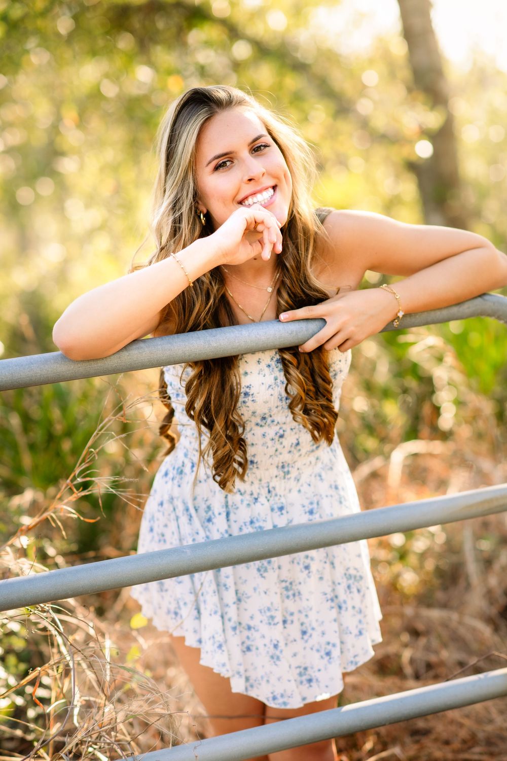 A person in a floral summer dress leans on a metal railing outdoors with a natural bokeh background.