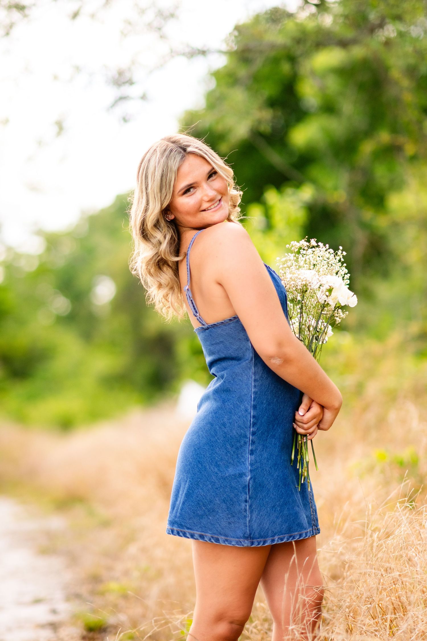 A person in a blue denim dress holds a bouquet of white wildflowers while standing in a sunlit field.