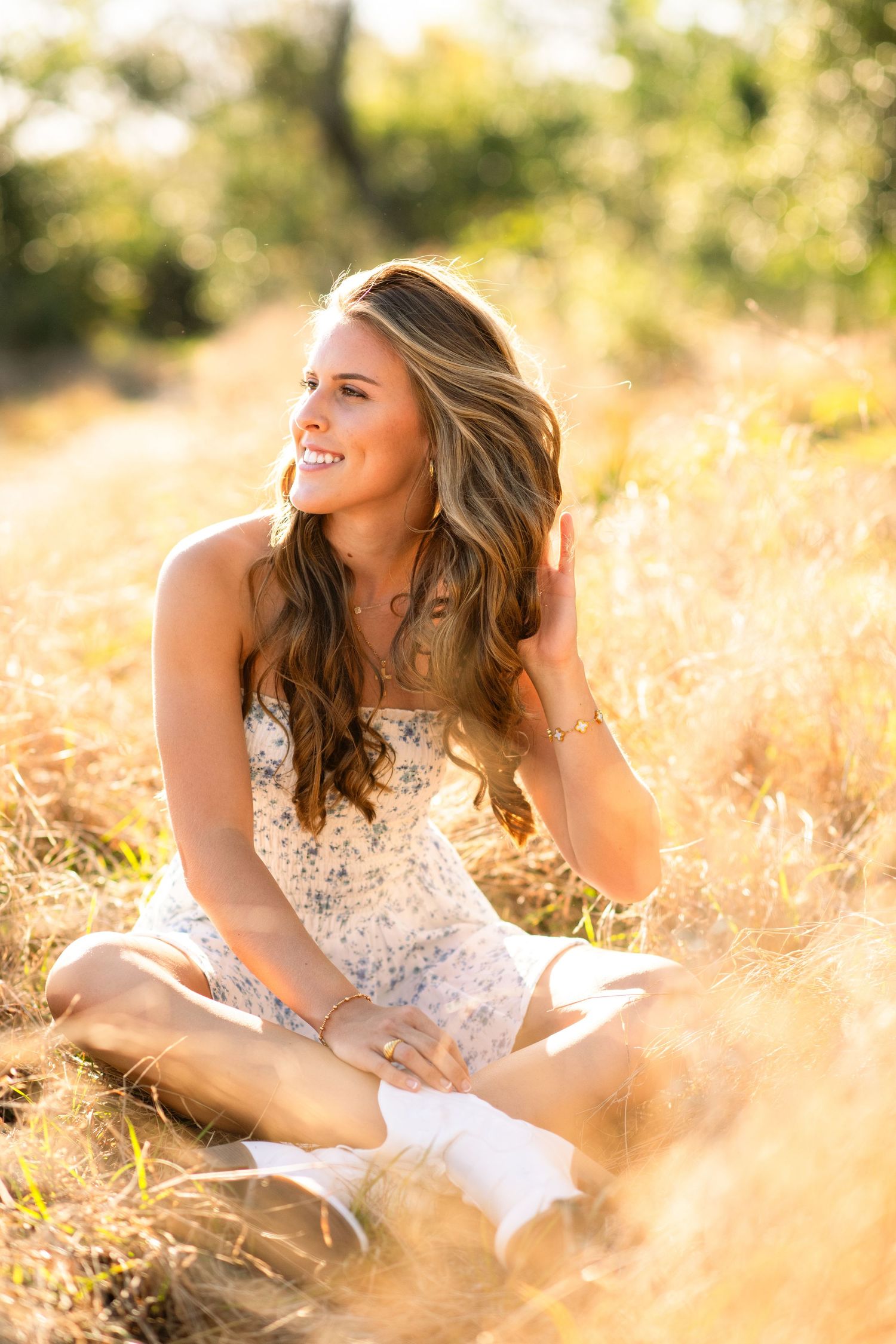A person in a white lace dress sits cross-legged in a sunlit field of golden grass during golden hour.