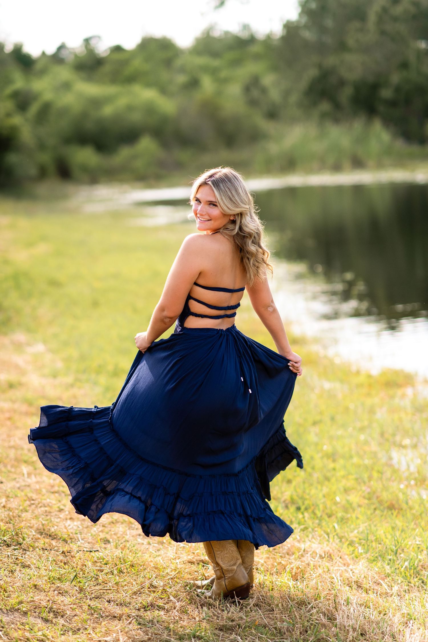A flowing navy blue dress twirls in motion near a serene lake on a sunny day, paired with cowboy boots.