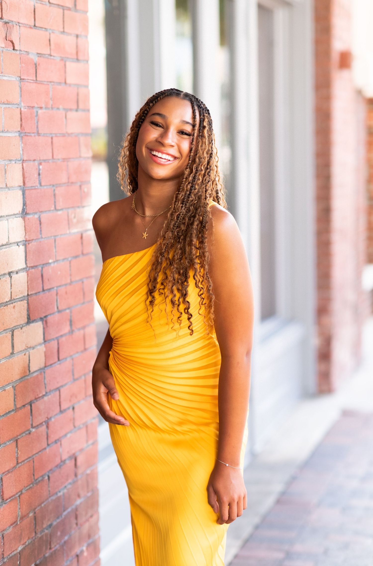 Person in vibrant yellow dress stands smiling against urban brick wall background.