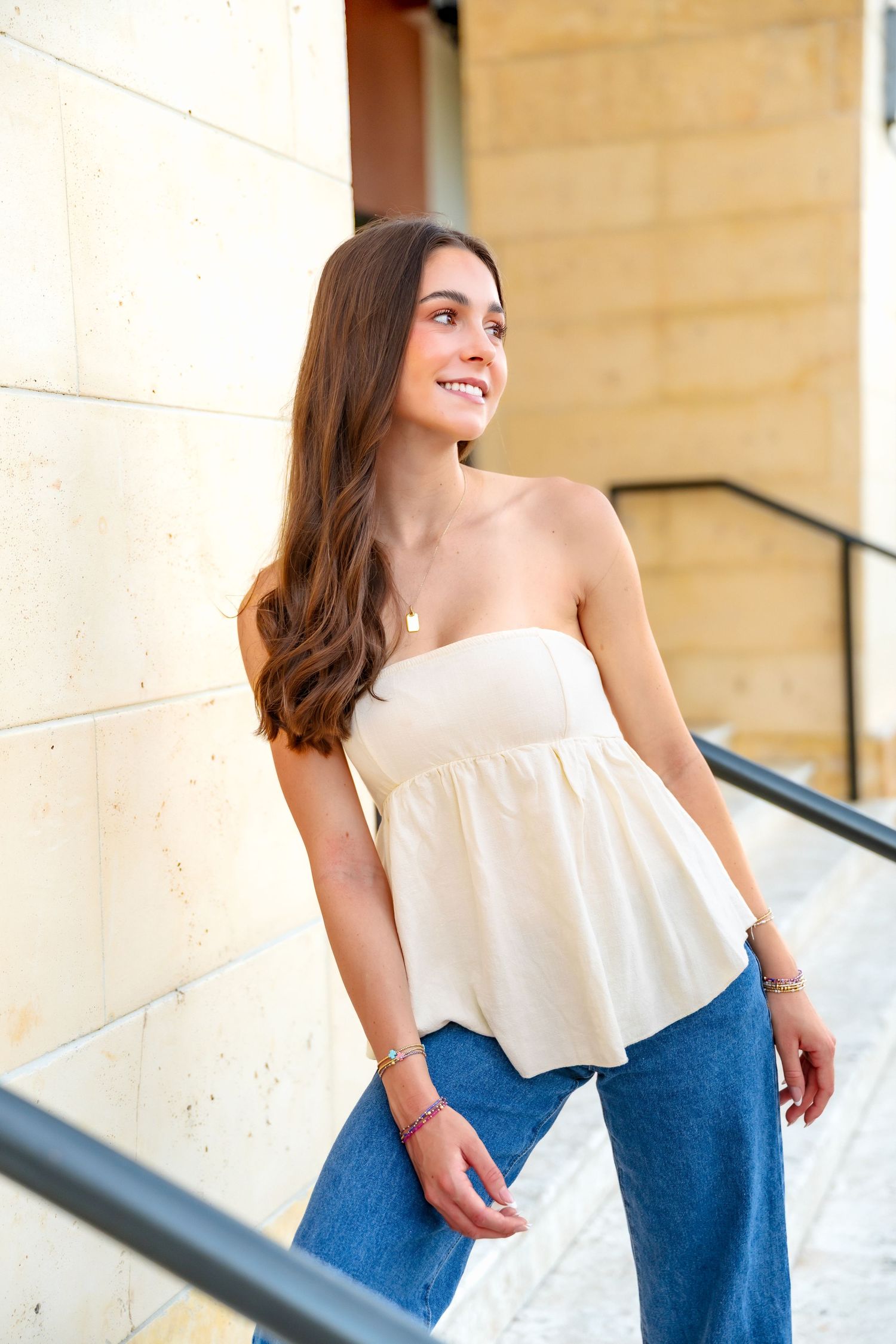 Young woman smiling in a cream strapless peplum top and blue wide-leg jeans, posing on outdoor steps.