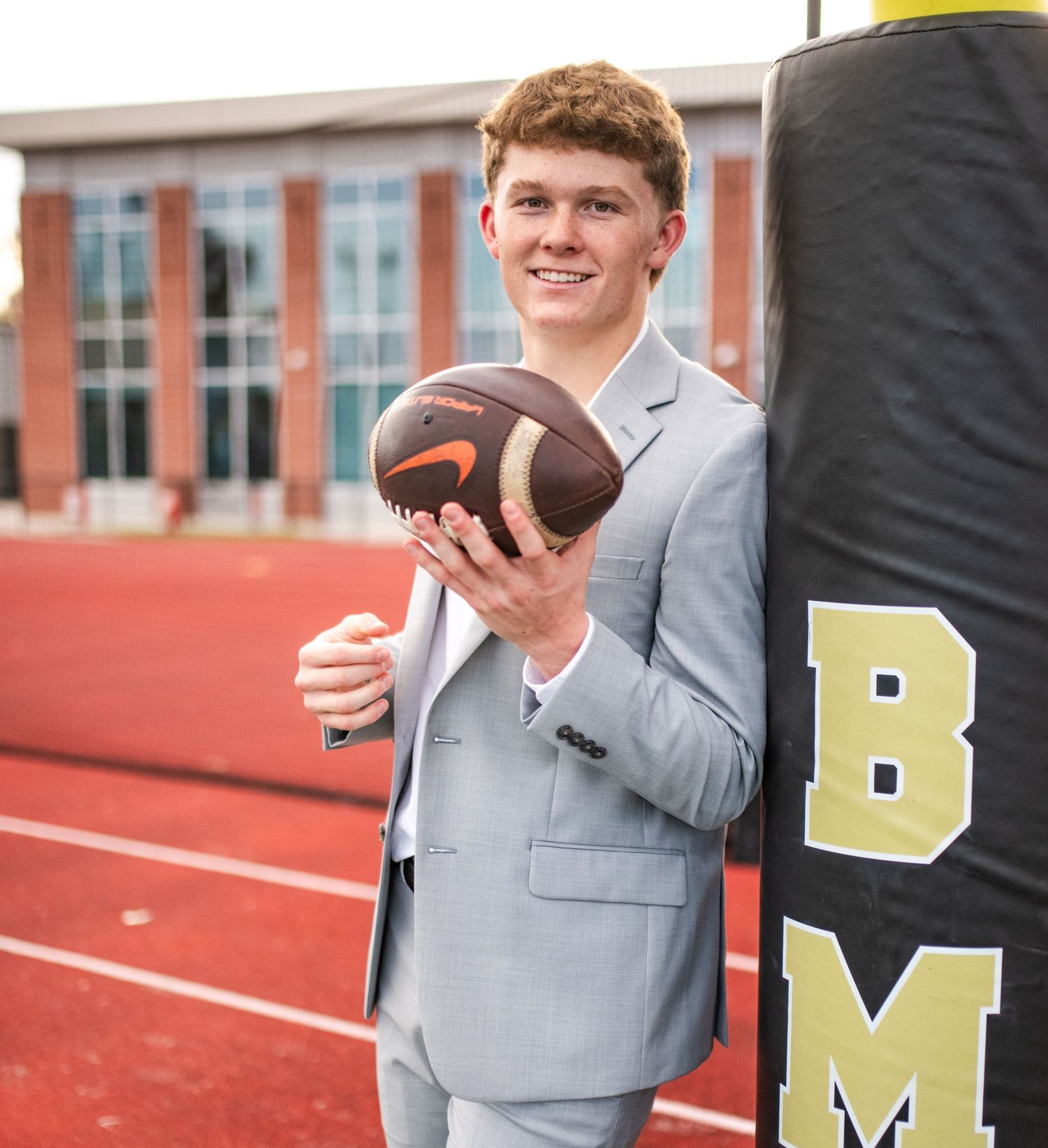 Student in gray suit and white shirt stands confidently in front of school sports bleachers.