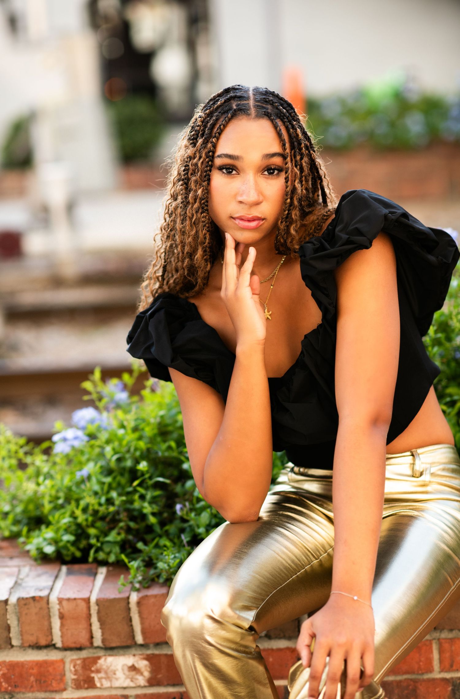 Stylish young woman in black ruffled top and metallic gold pants poses on brick steps surrounded by greenery.