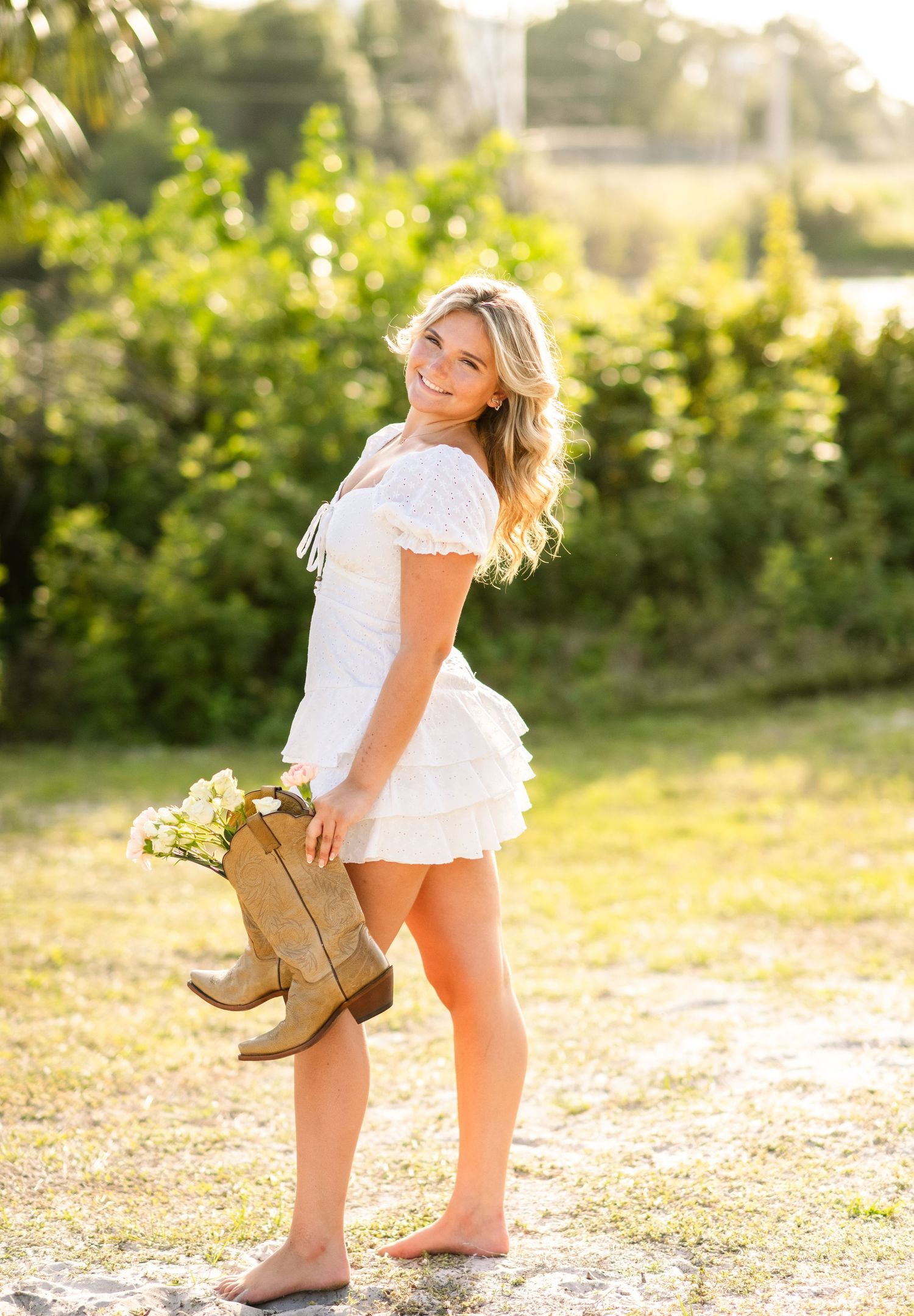 A person in a white dress stands barefoot on a sandy ground against a sunlit green foliage background.
