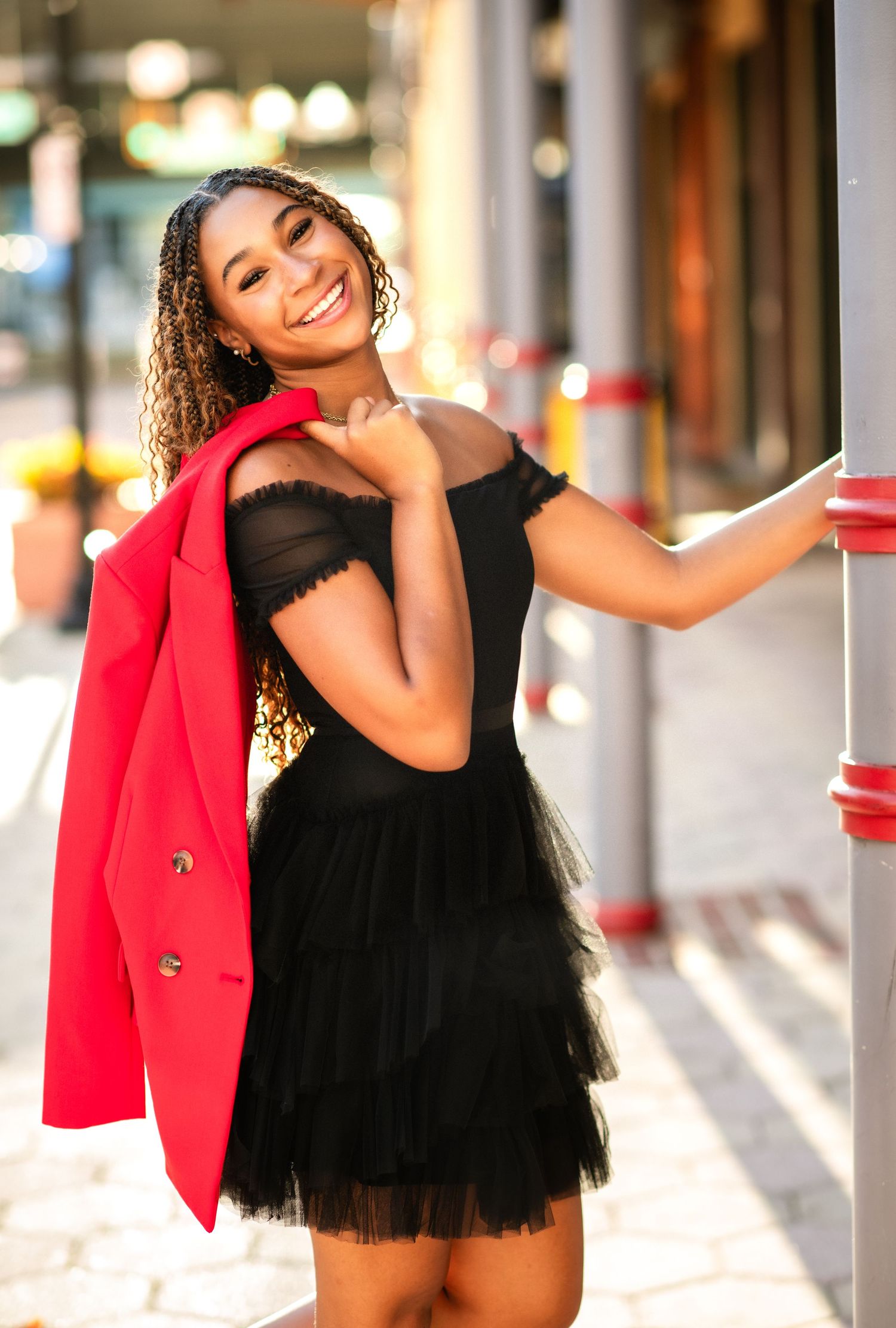 Fashion model in black mini dress and red blazer poses confidently in bright urban setting.