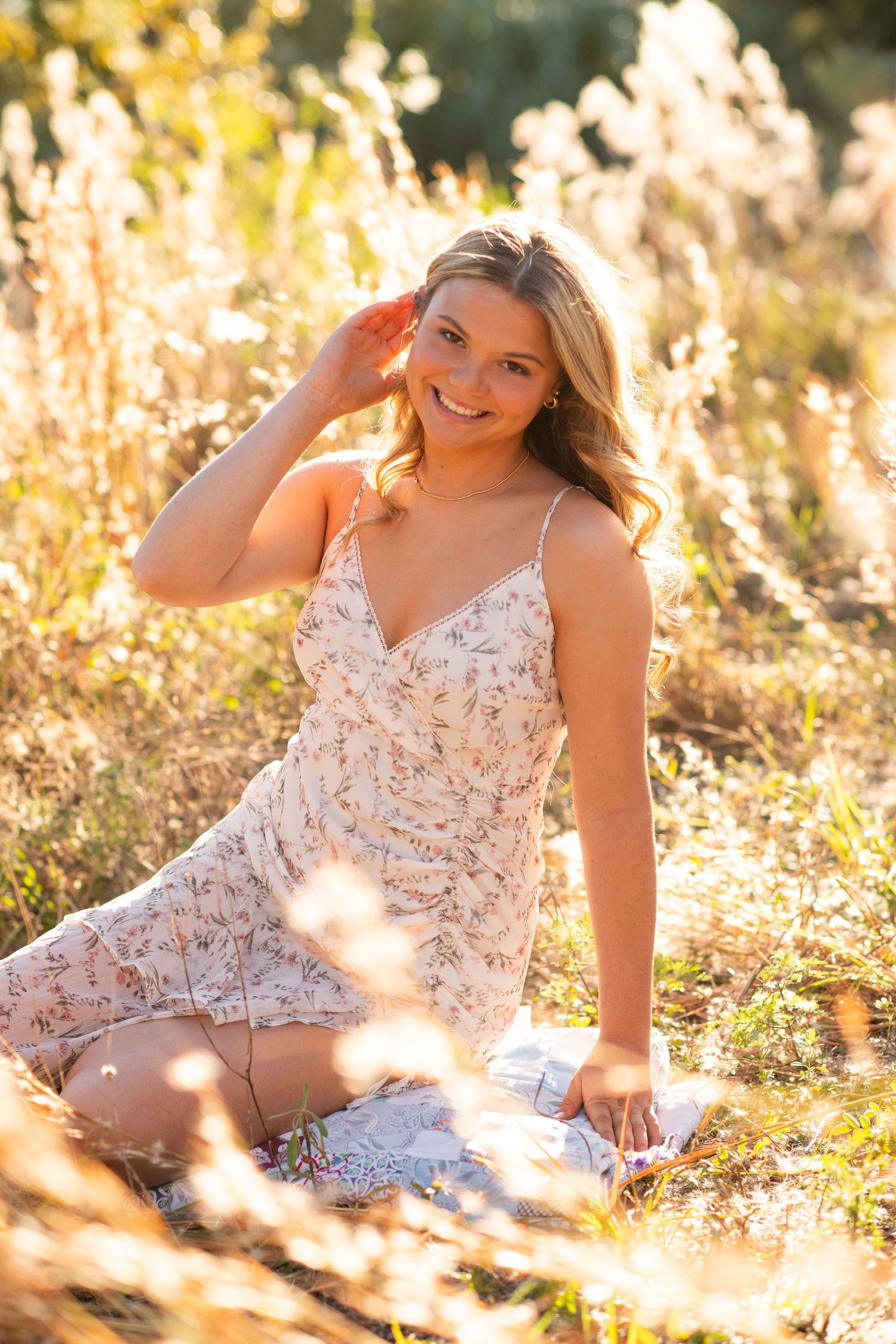 A sunlit outdoor photoshoot featuring someone in a floral dress laying in golden grasses during sunset.