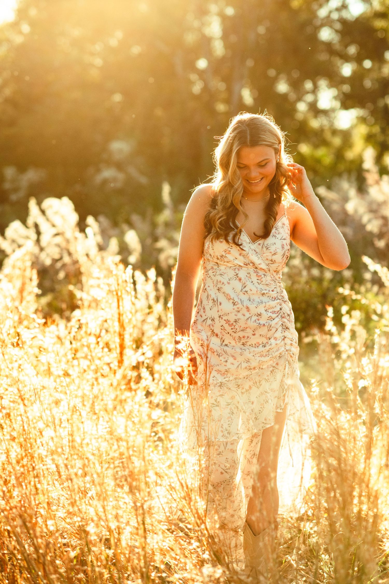 Woman in white sleeveless dress stands thoughtfully in golden sunlit field of tall grasses at sunset.