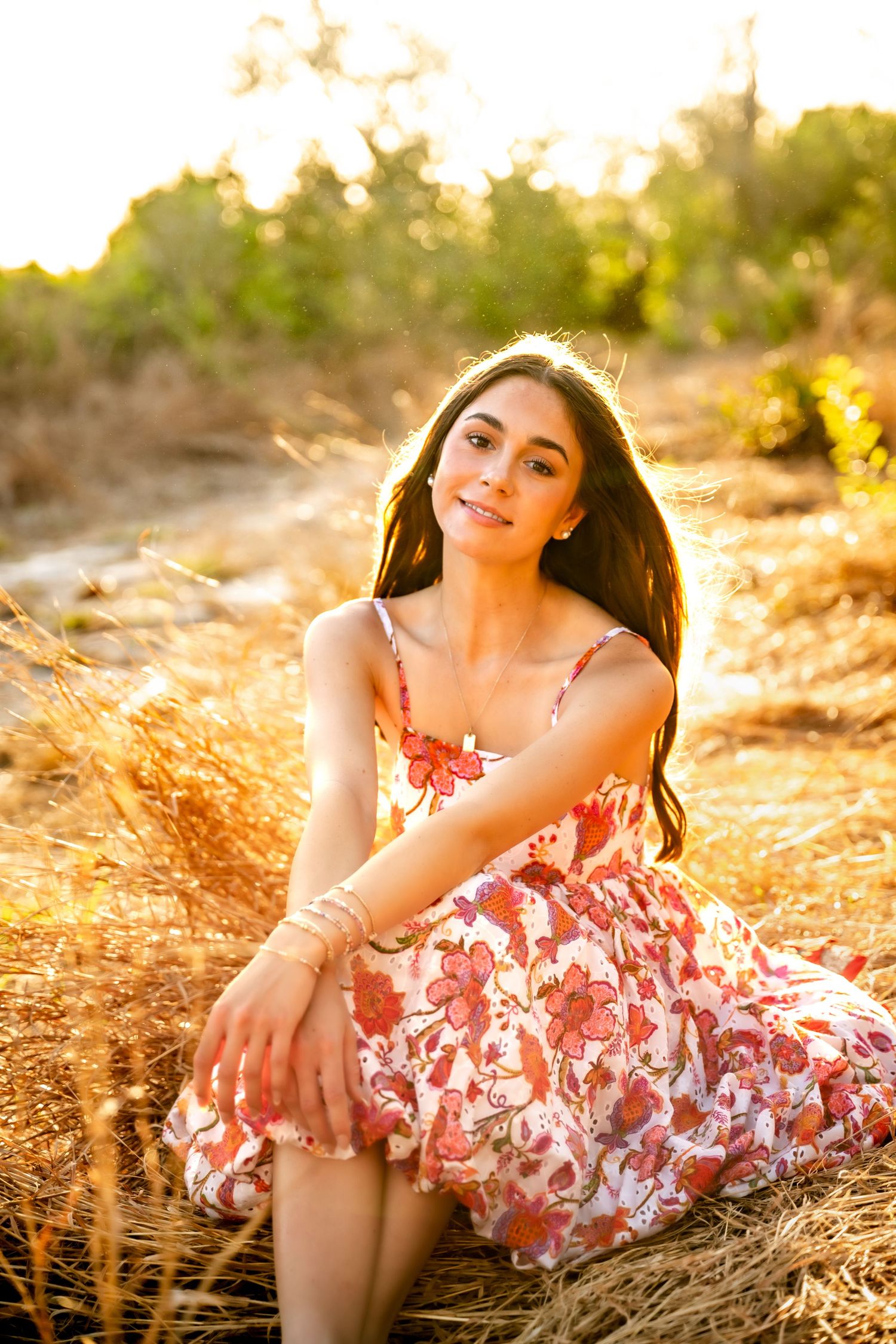 Young woman in floral dress sitting in golden field at sunset, smiling softly with long dark hair.