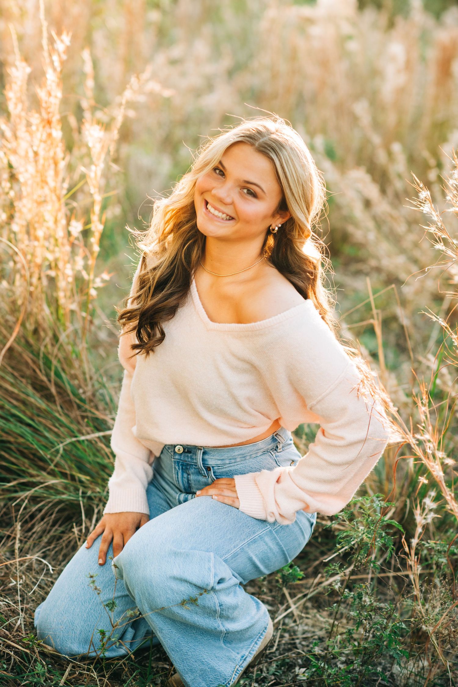 Blonde woman in white sweater and jeans sits in golden grass field at sunset with warm lighting.