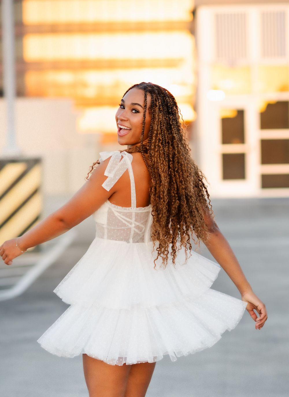 A dancer in a white tulle mini dress with corset bodice twirls joyfully in an urban setting at sunset.
