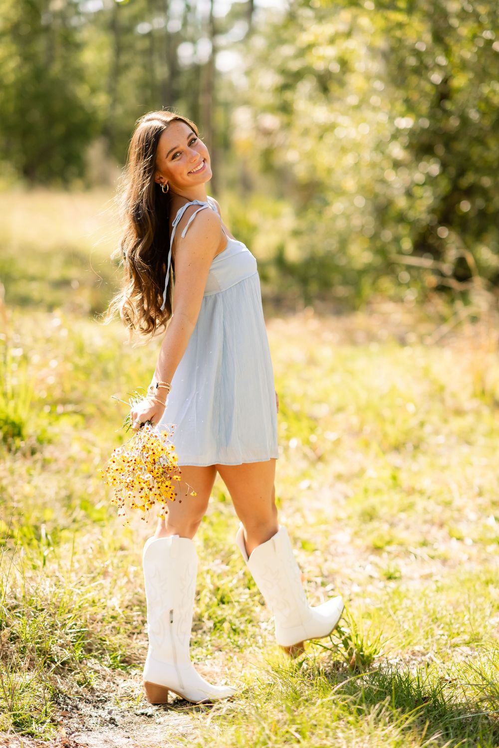 A person in a light-colored dress and white boots poses with wildflowers in a sun-drenched meadow on a summer day.