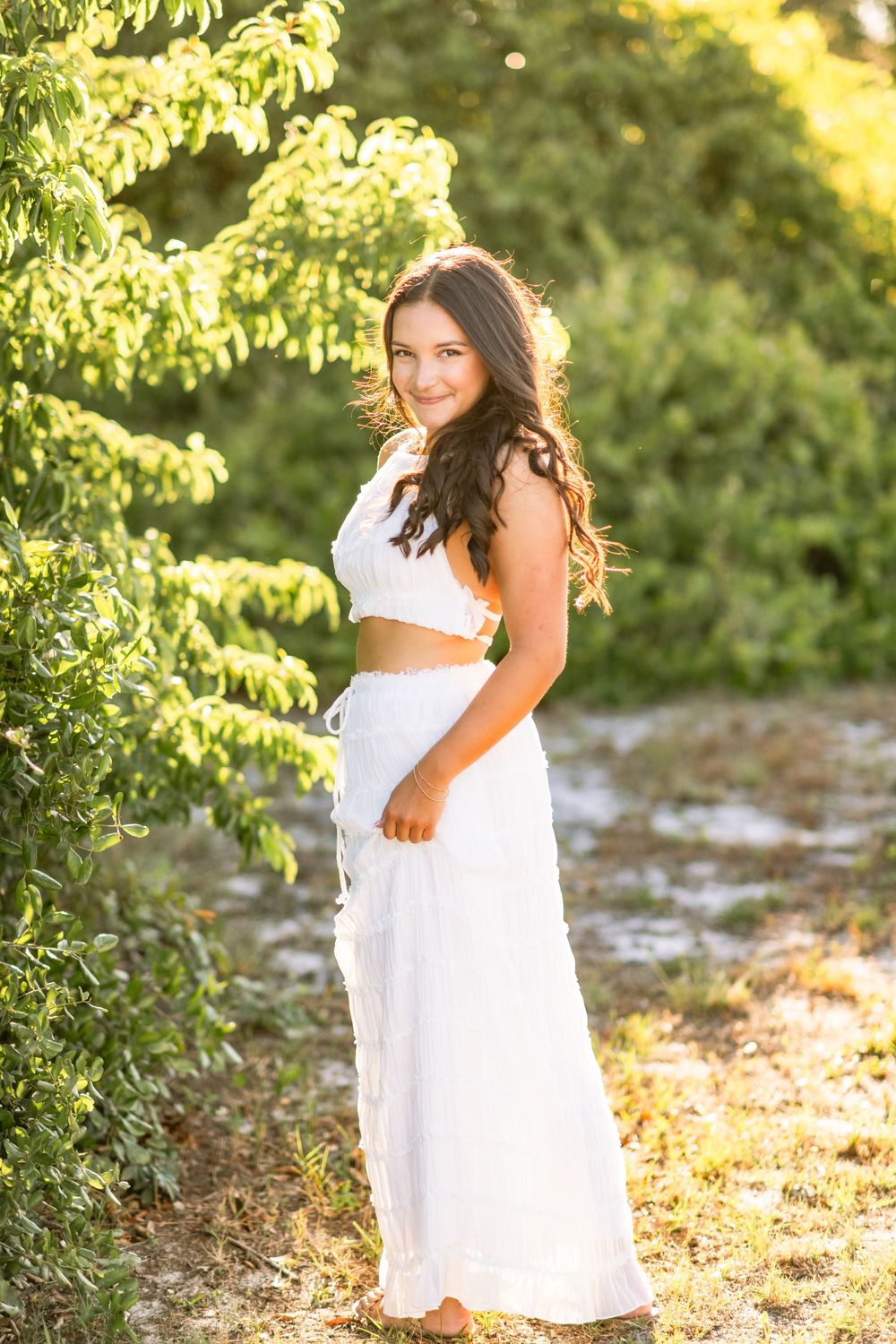 A dreamy golden hour portrait capturing someone in a white two-piece outfit standing beside lush green foliage.