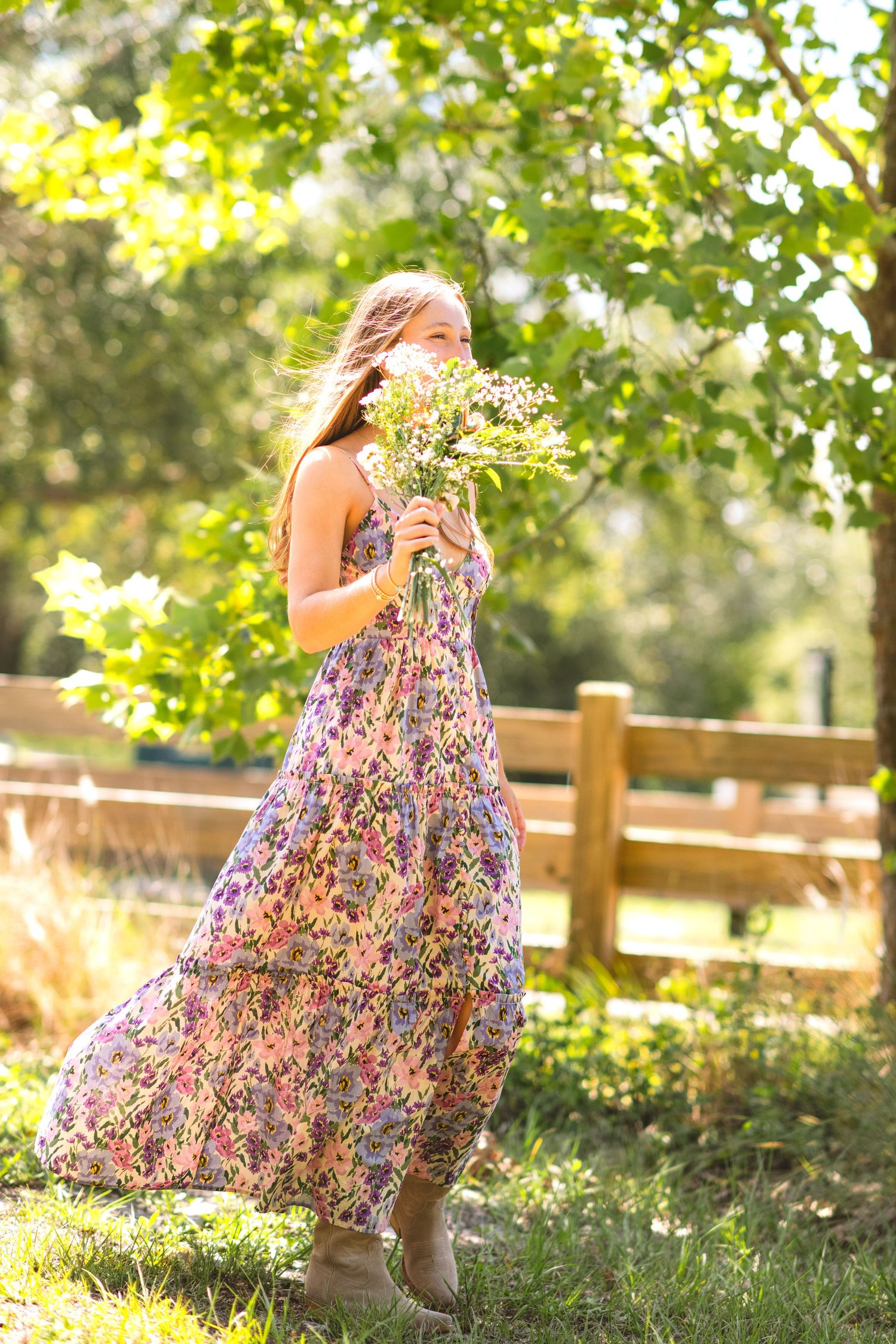 Girl in floral dress twirls and plays in a sunlit garden with wooden fence and greenery in the background.