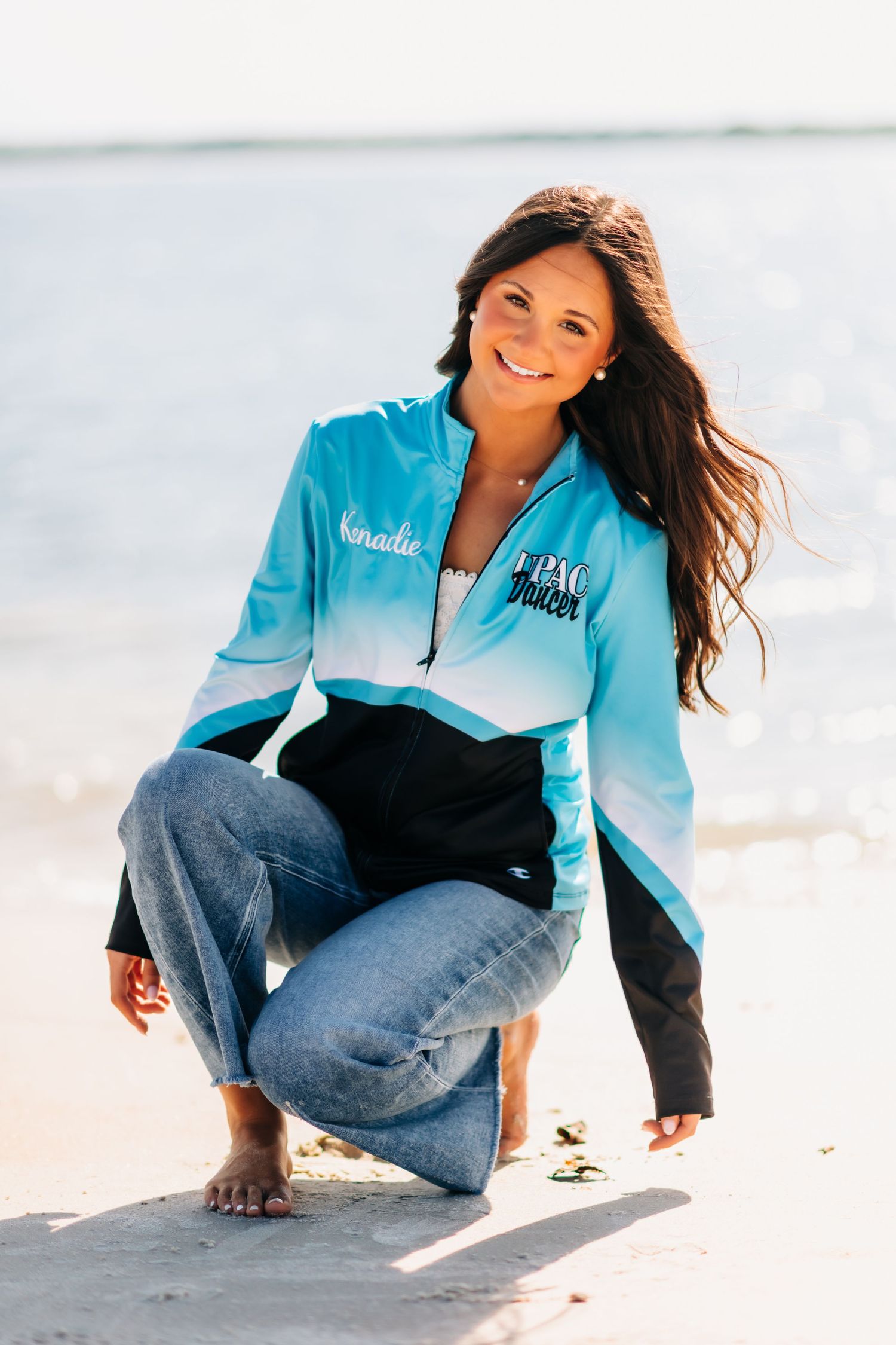 Woman in teal ombre zip-up jacket and jeans crouching barefoot on sandy beach, smiling with long dark hair.