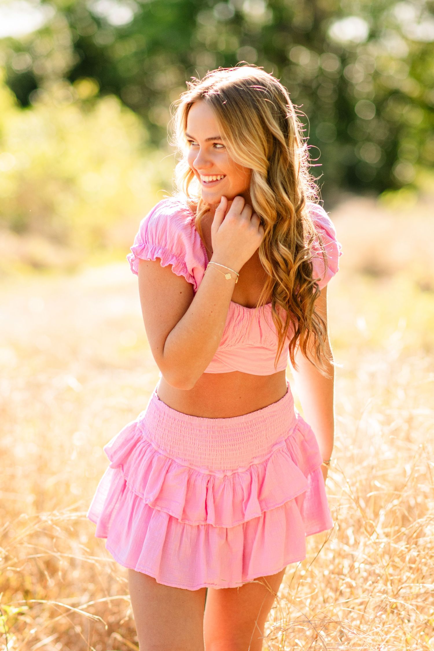 Woman in pink ruffled dress laughs and dances in a golden sunlit meadow field during sunset.