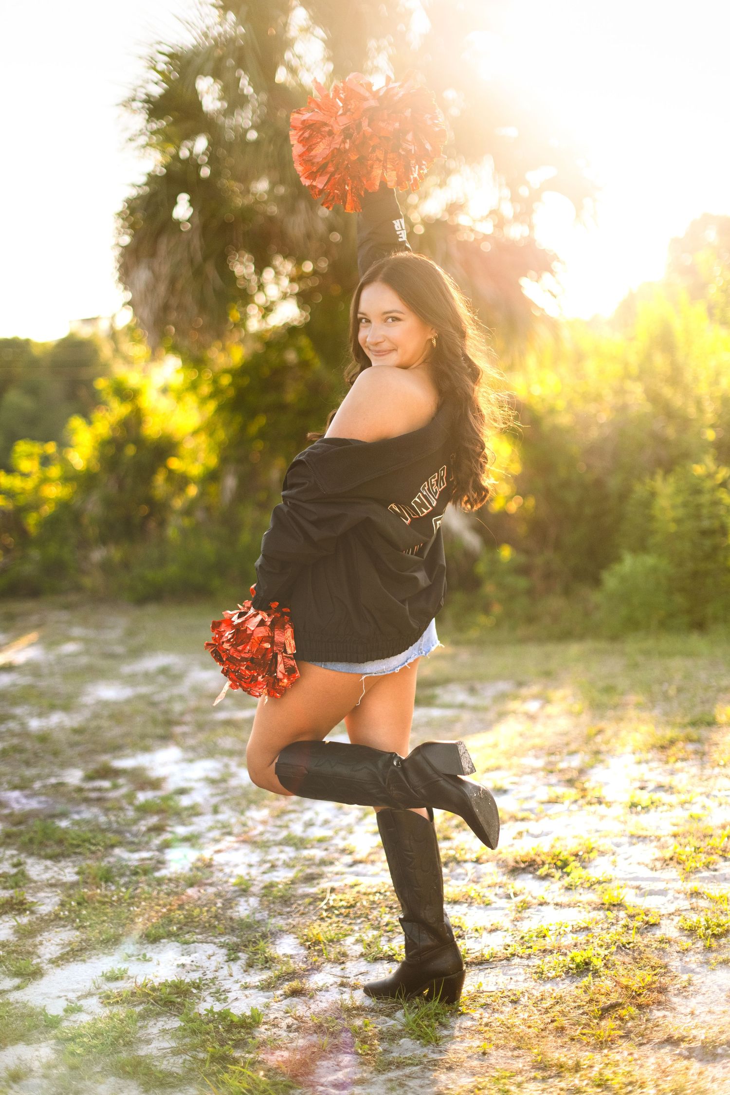 Woman in black sweater and tall boots poses outdoors during golden hour sunset with pom poms in natural setting.