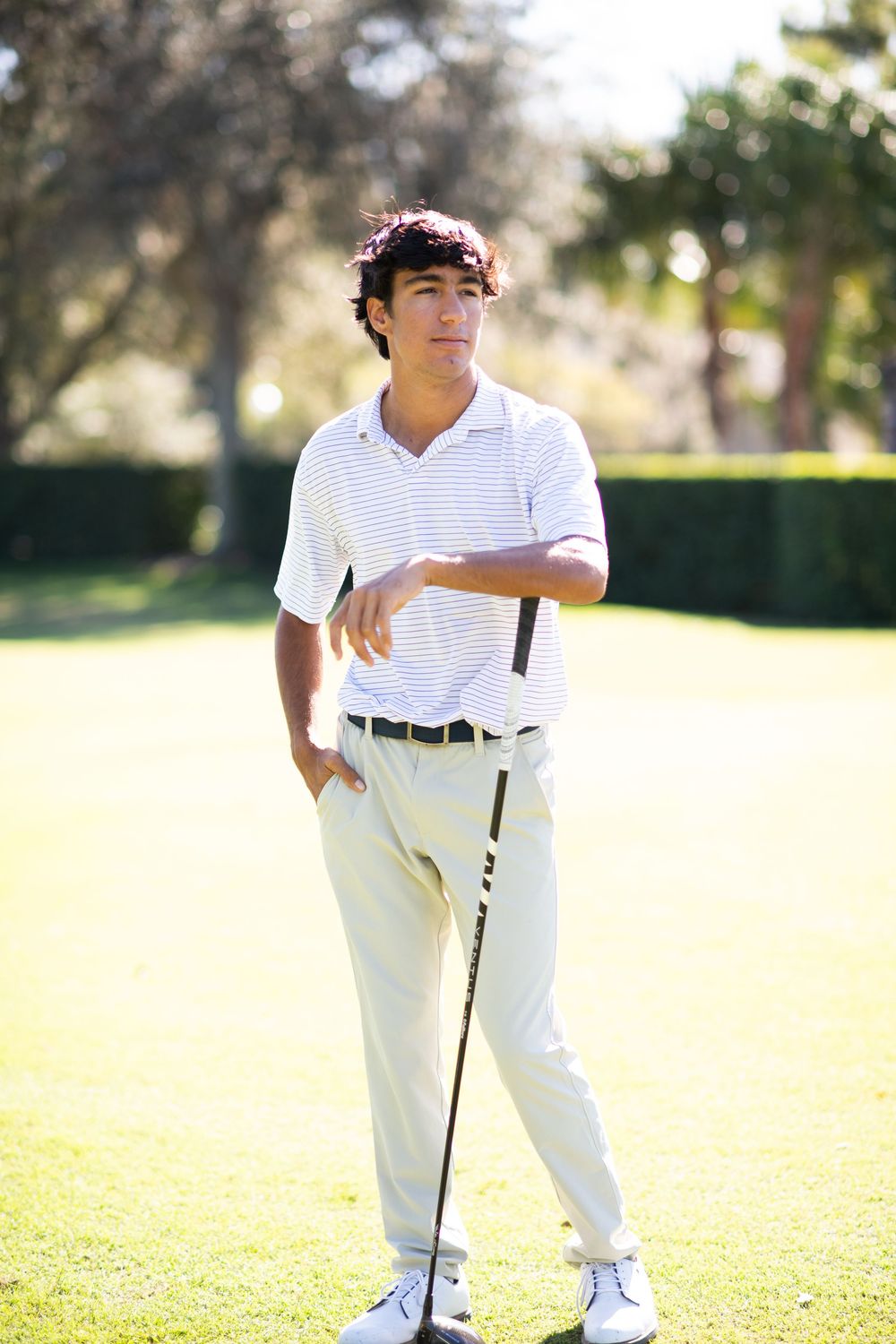 Young golfer in all-white attire stands casually with golf club on sunny course during summer day.