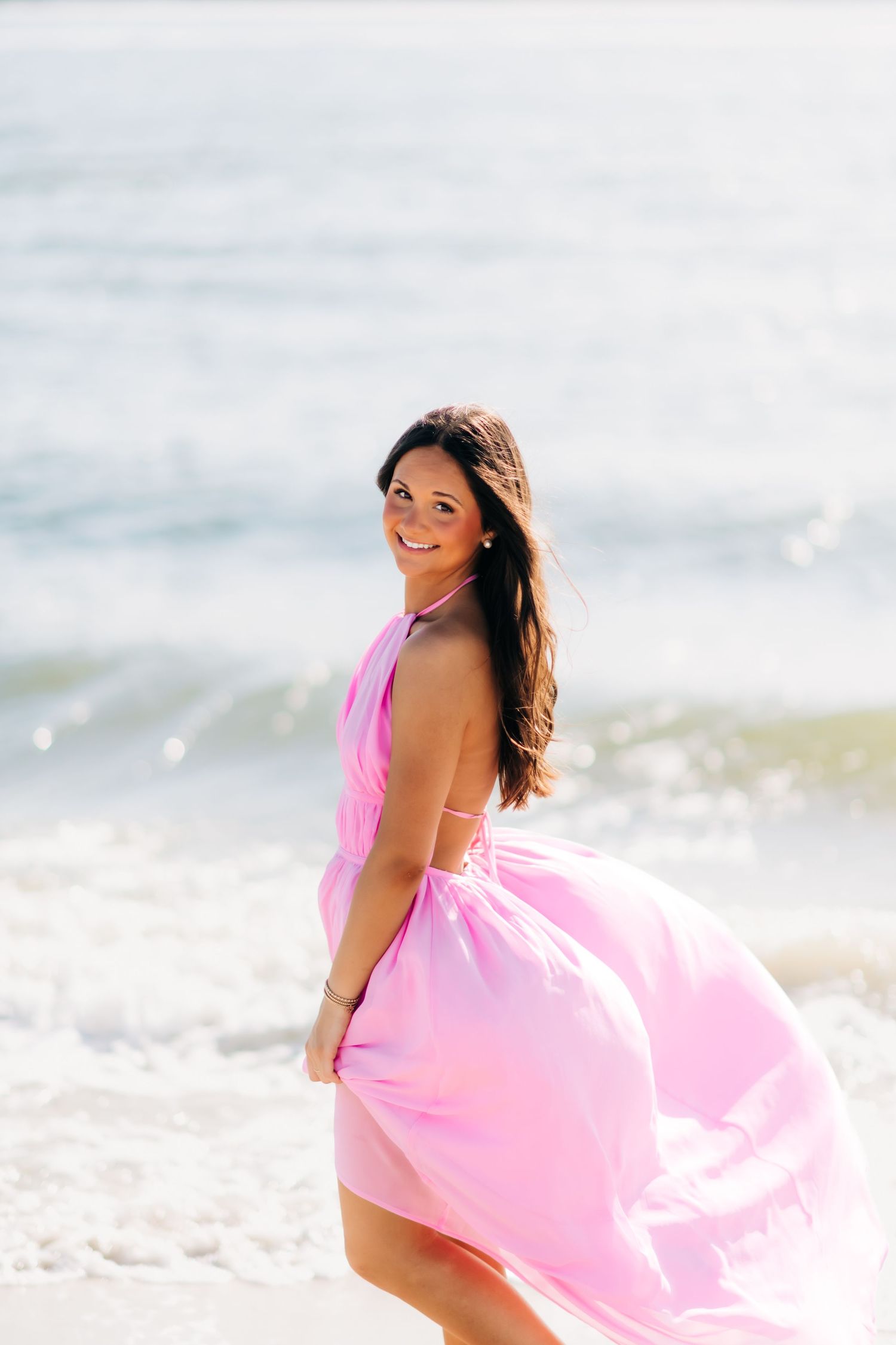 Smiling brunette woman in flowing pink halter dress on sunny beach with ocean waves in background.