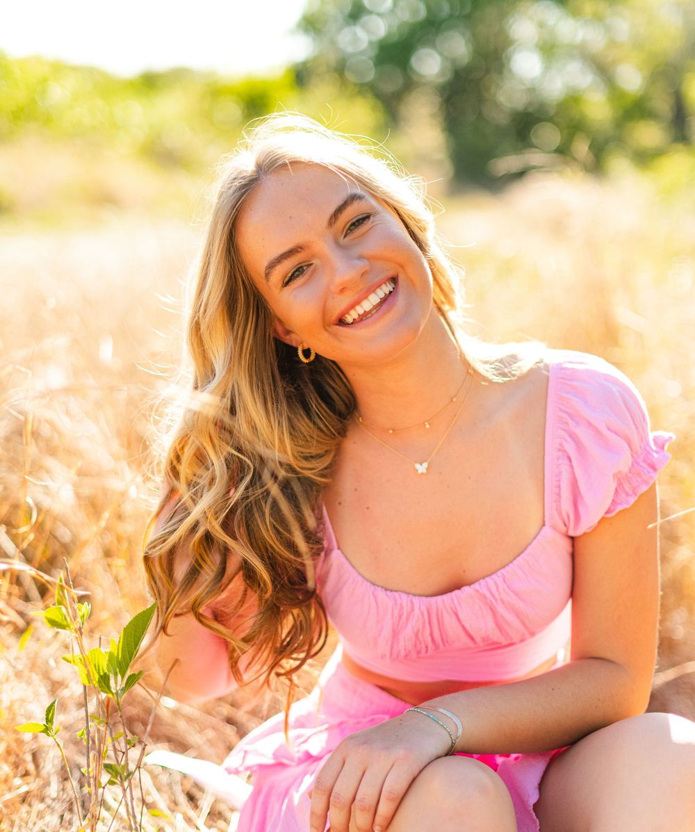 Person in pink summer dress smiles while sitting in golden sunlit field of tall grass on warm afternoon.