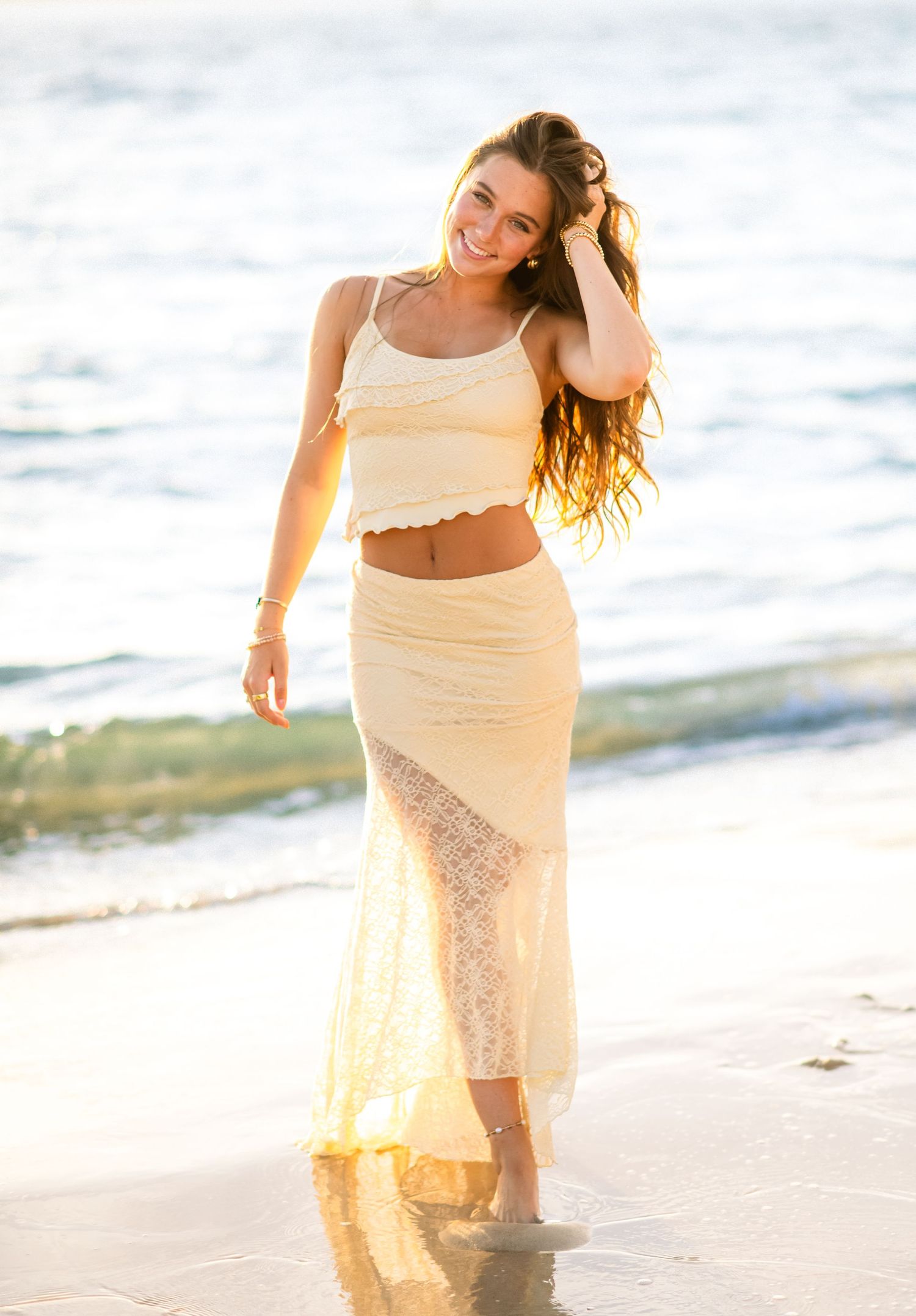 Person in cream-colored two-piece outfit walking on sunny beach shore at sunset with waves in background.