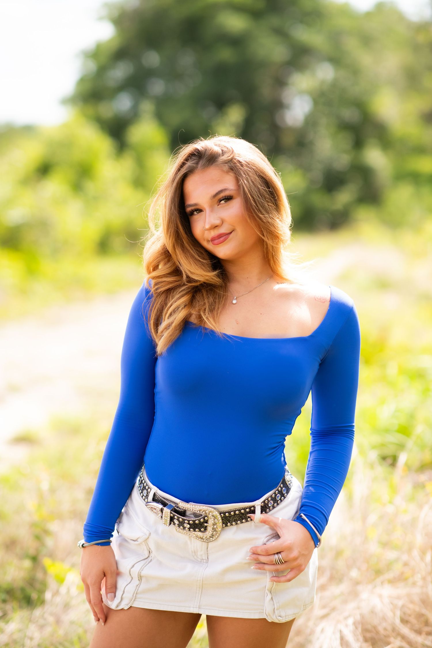 Person wearing a bright royal blue long-sleeved top and white shorts poses in a sunny outdoor field setting.