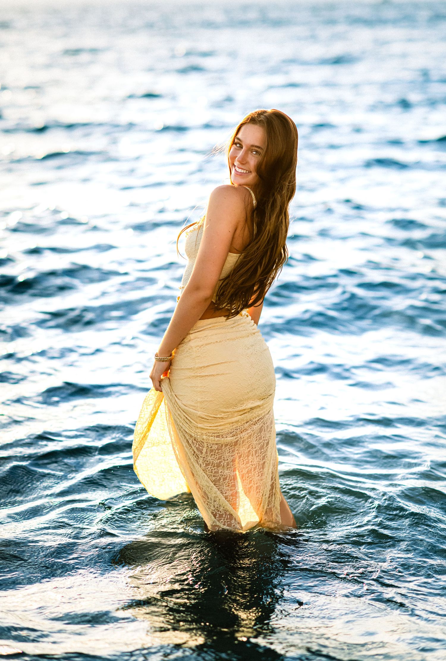 A person wearing a sheer yellow dress stands in ocean waves at sunset, creating a dreamy silhouette against the water.