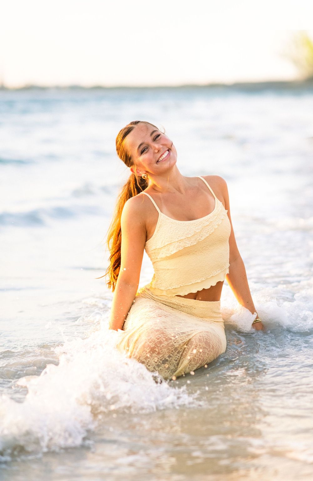Someone in a cream-colored dress joyfully playing in ocean waves during golden hour as sunlight creates a dreamy atmosphere.