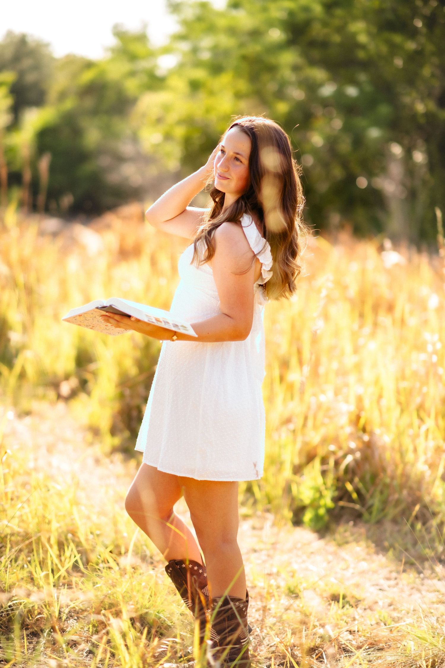 Young woman in white dress and cowboy boots holding a book in a sunlit golden field.