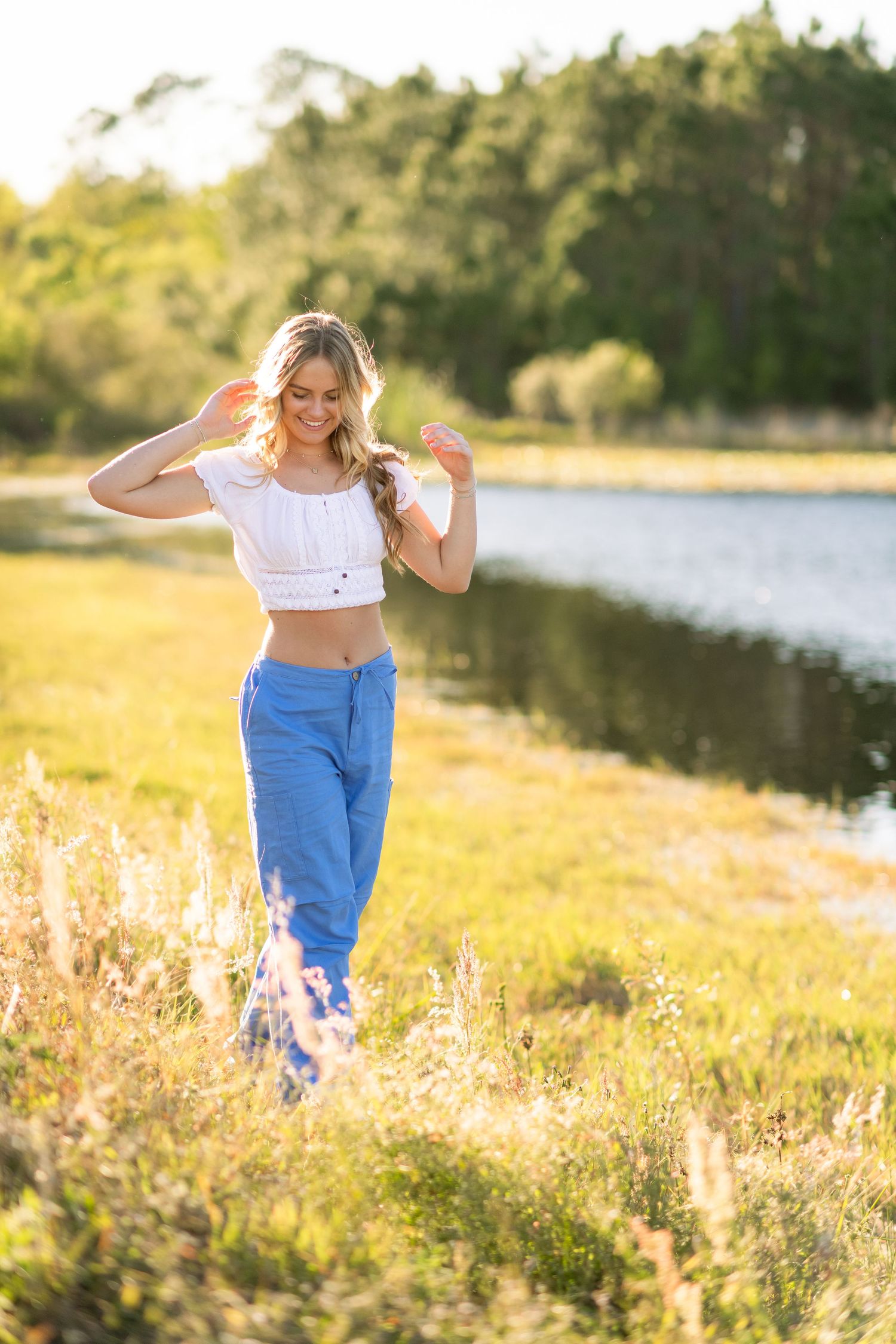 Young woman in white crop top and blue pants dancing in a sunlit meadow by a lake at golden hour.