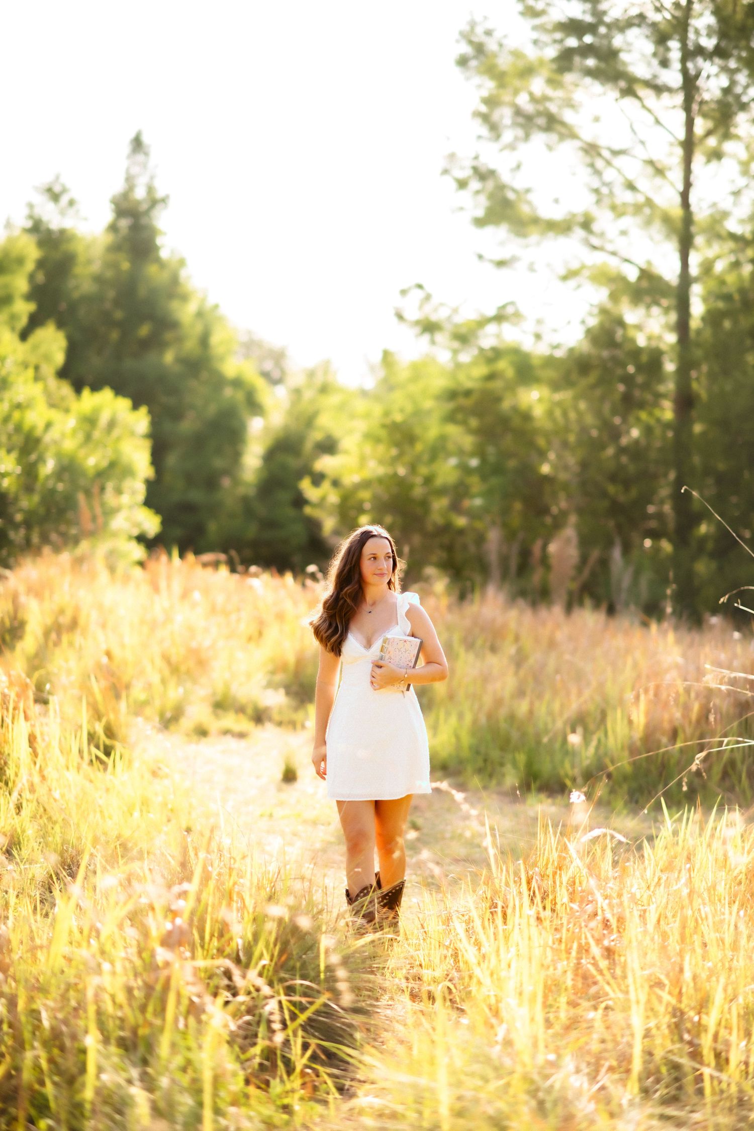Young woman in white dress and cowboy boots walks through golden meadow holding a book, bathed in warm sunlight.