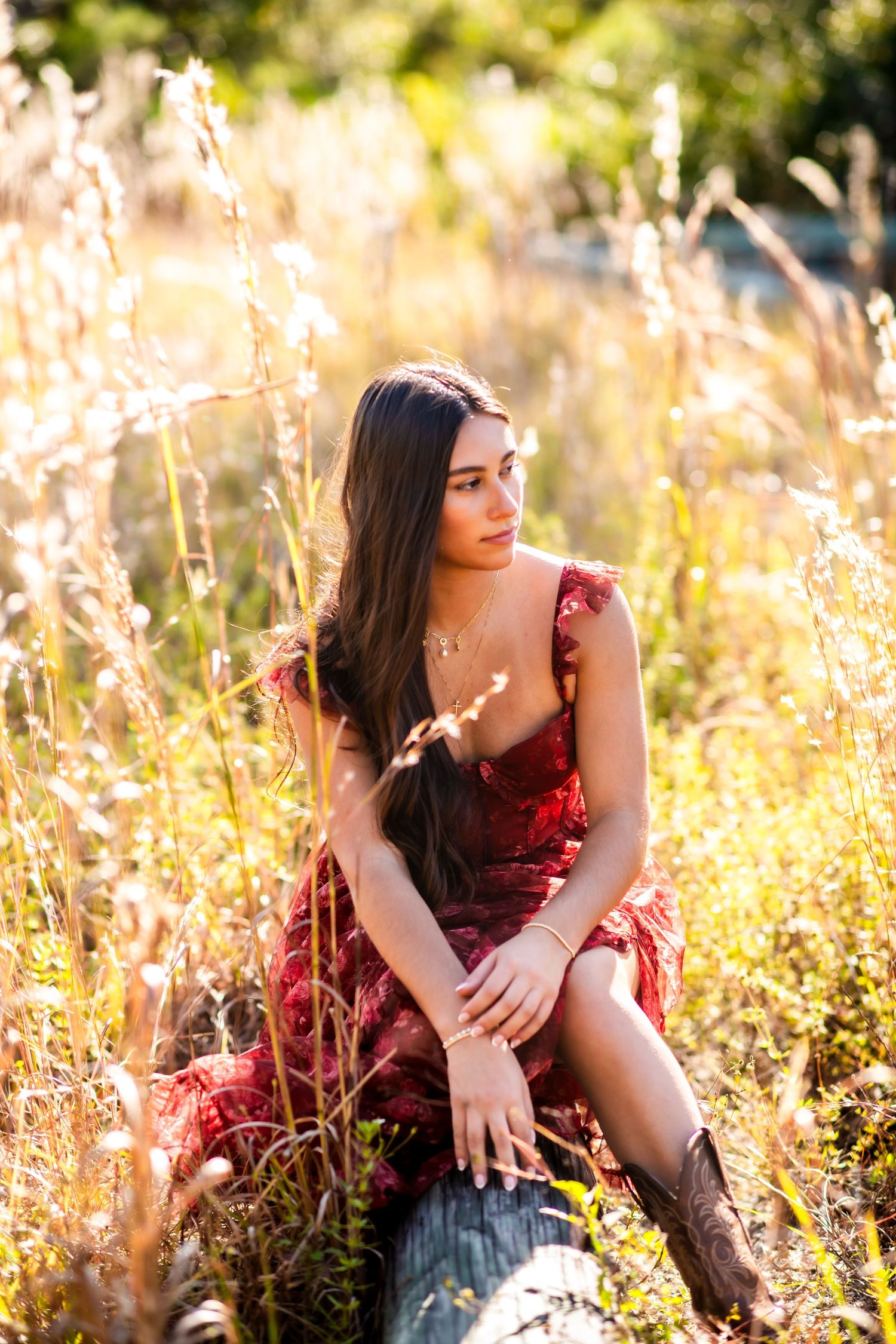 Young woman in red floral dress sitting in golden field grass during sunset.