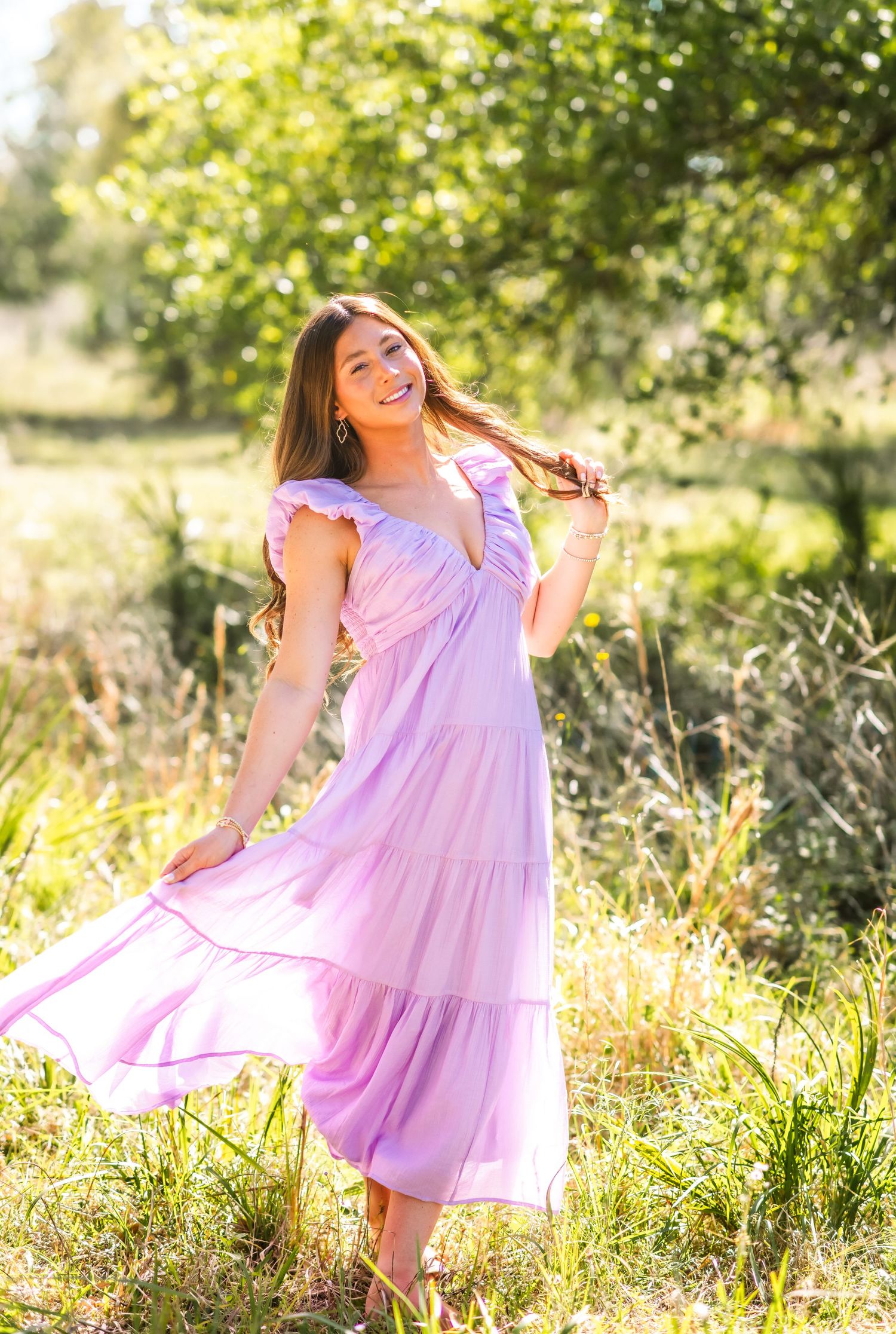 Light purple tiered maxi dress flows in a sunlit meadow with golden wildflowers and green trees.