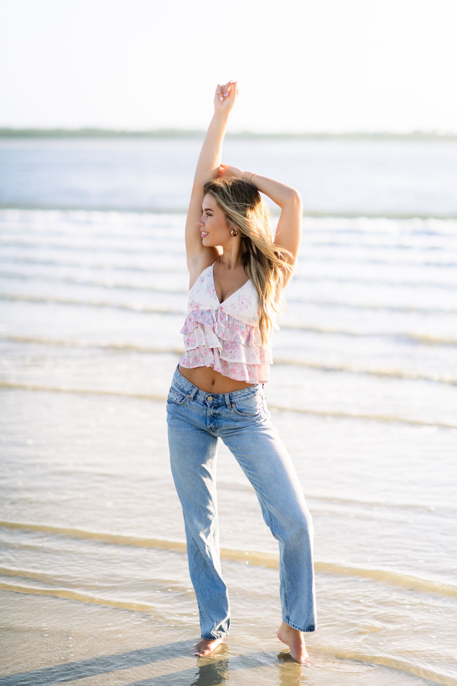 A woman dances joyfully in shallow water at the beach wearing a floral crop top and light blue jeans at sunset.