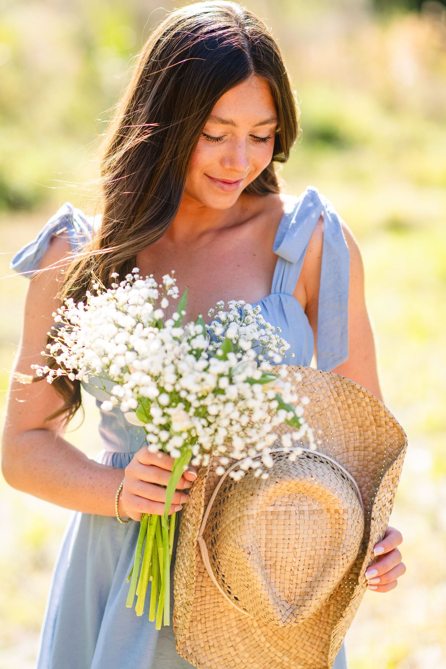 Person in a blue sundress holding a bouquet of white baby's breath flowers and straw hat in sunny outdoor setting.