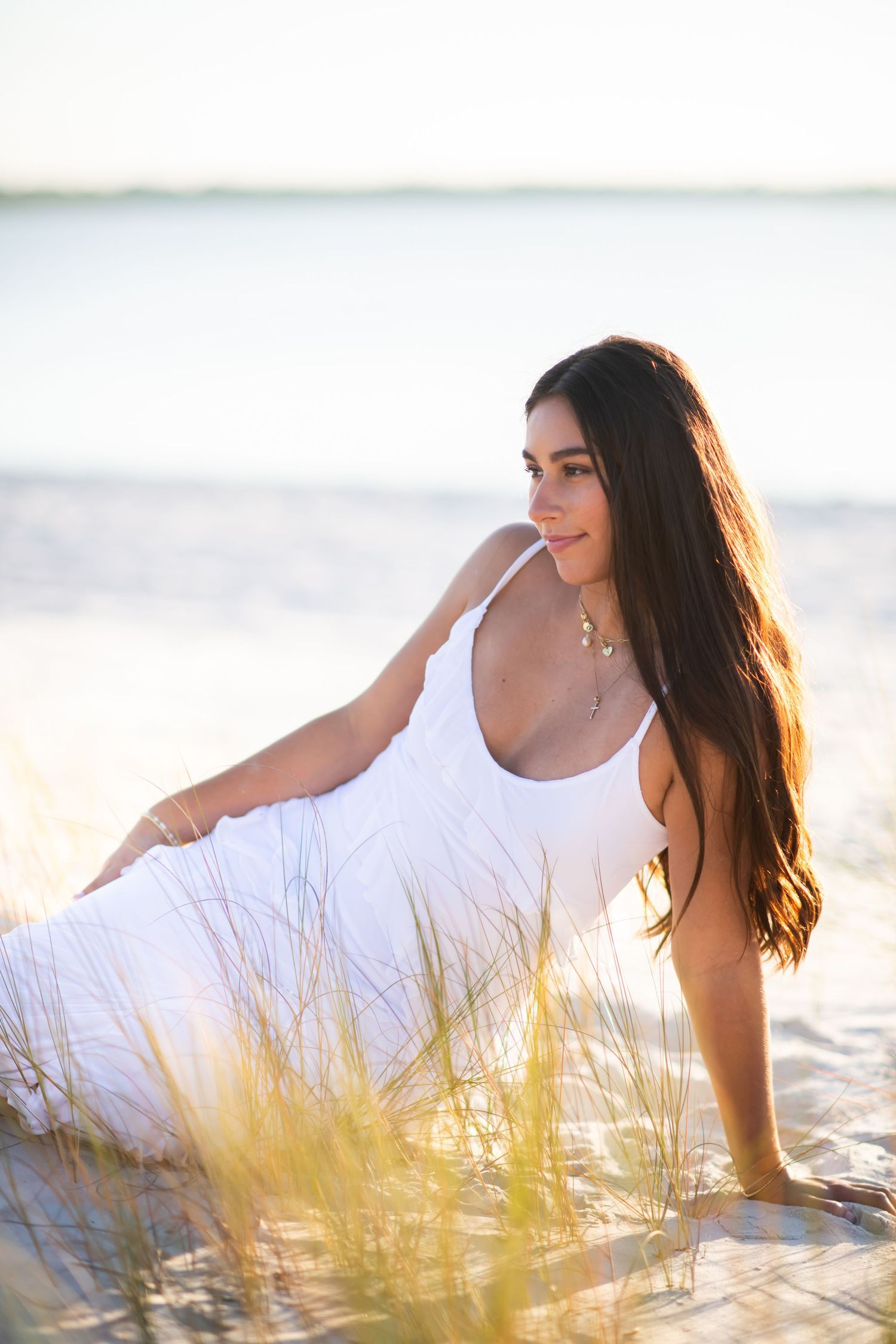Young woman in white sundress sitting on sandy beach dunes with long dark hair, golden hour sunlight, serene ocean backdrop.