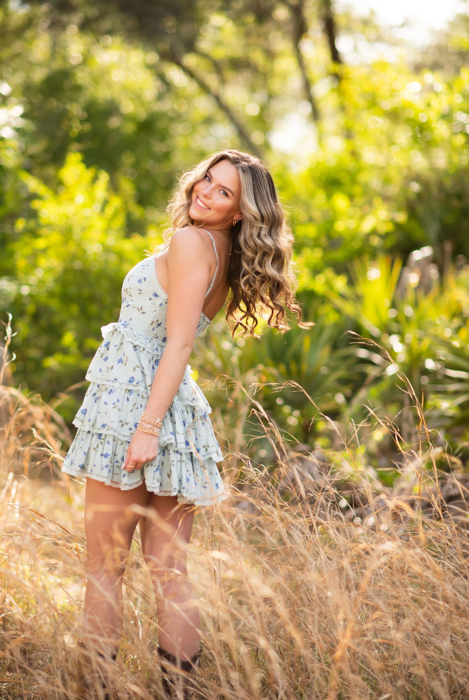A person in a light floral dress poses in tall grass during golden hour with sunlit trees in the background.