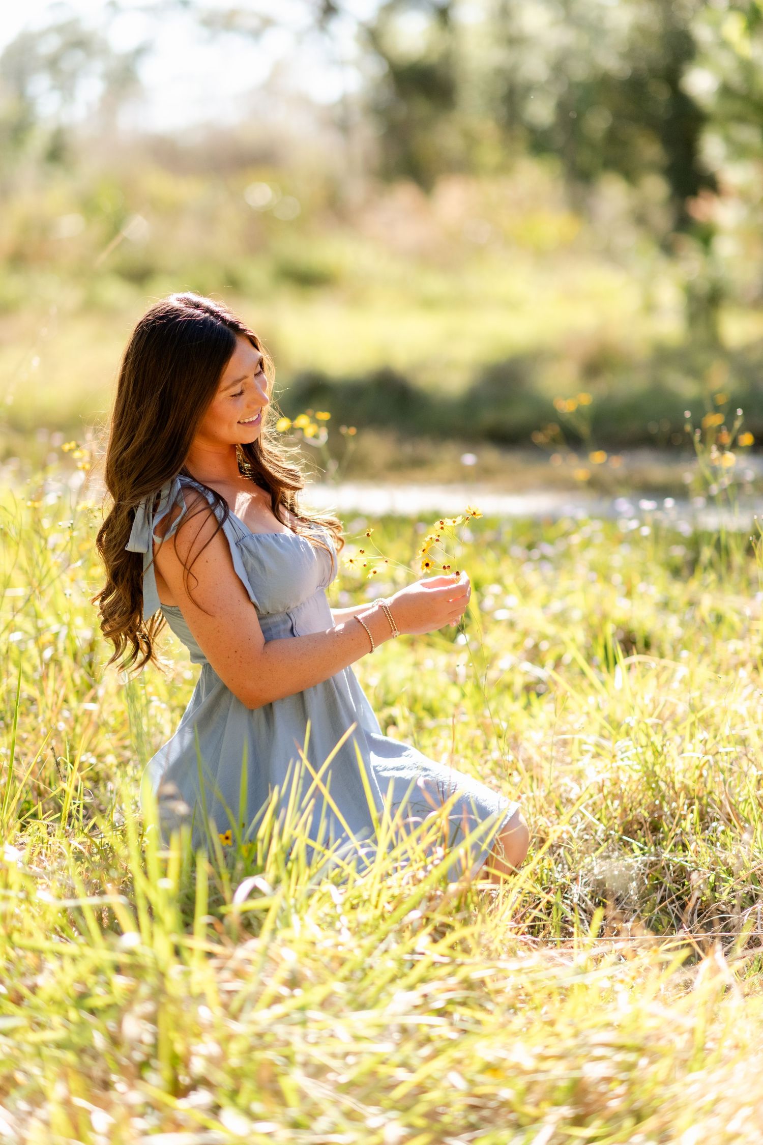 A person in a soft gray dress sits gracefully in a sun-dappled meadow of golden wildflowers on a bright afternoon.