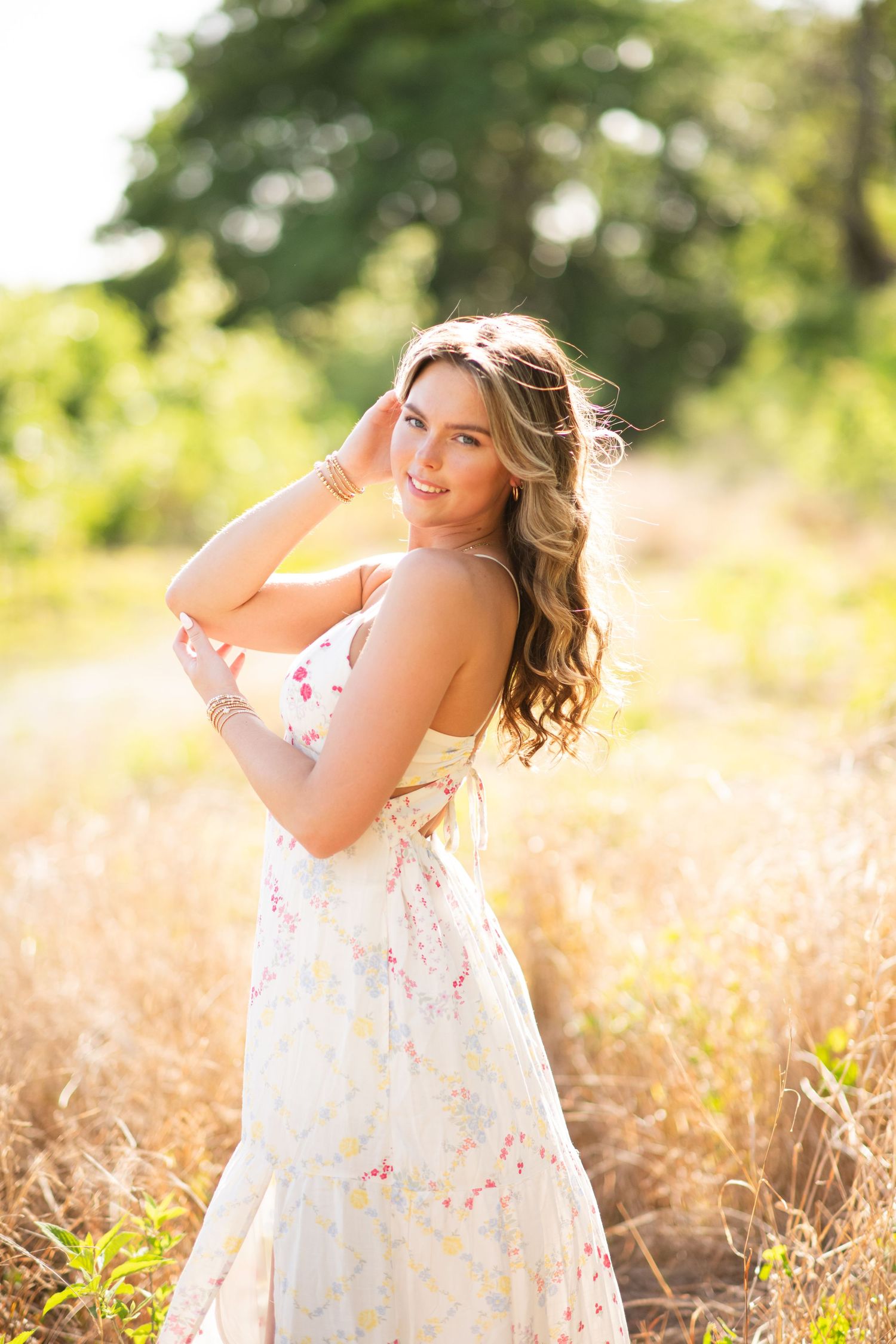 Woman in white floral sundress poses gracefully in a golden sunlit field during sunset photoshoot.